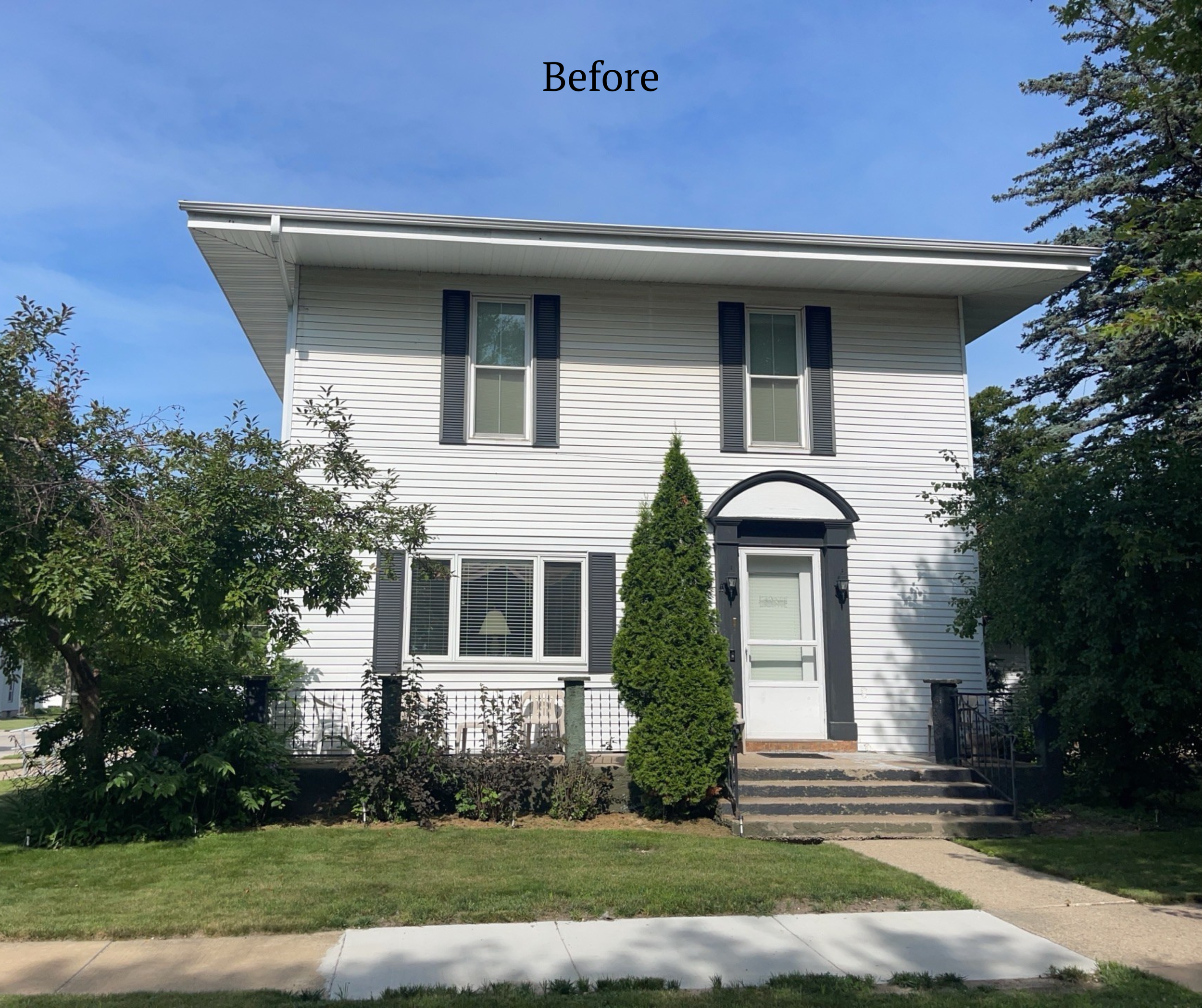 White two-story house with black shutters, a small front porch, and a tree in the front yard on a sunny day with a blue sky.