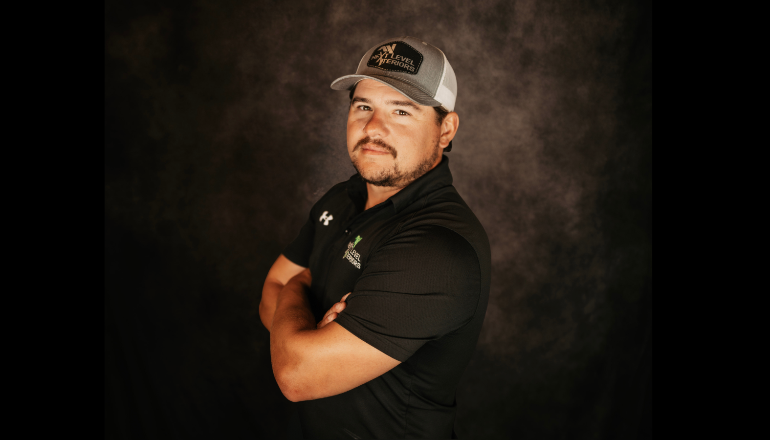 A smiling man with a beard, wearing a baseball cap and a black polo shirt with logos, standing against a dark backdrop.