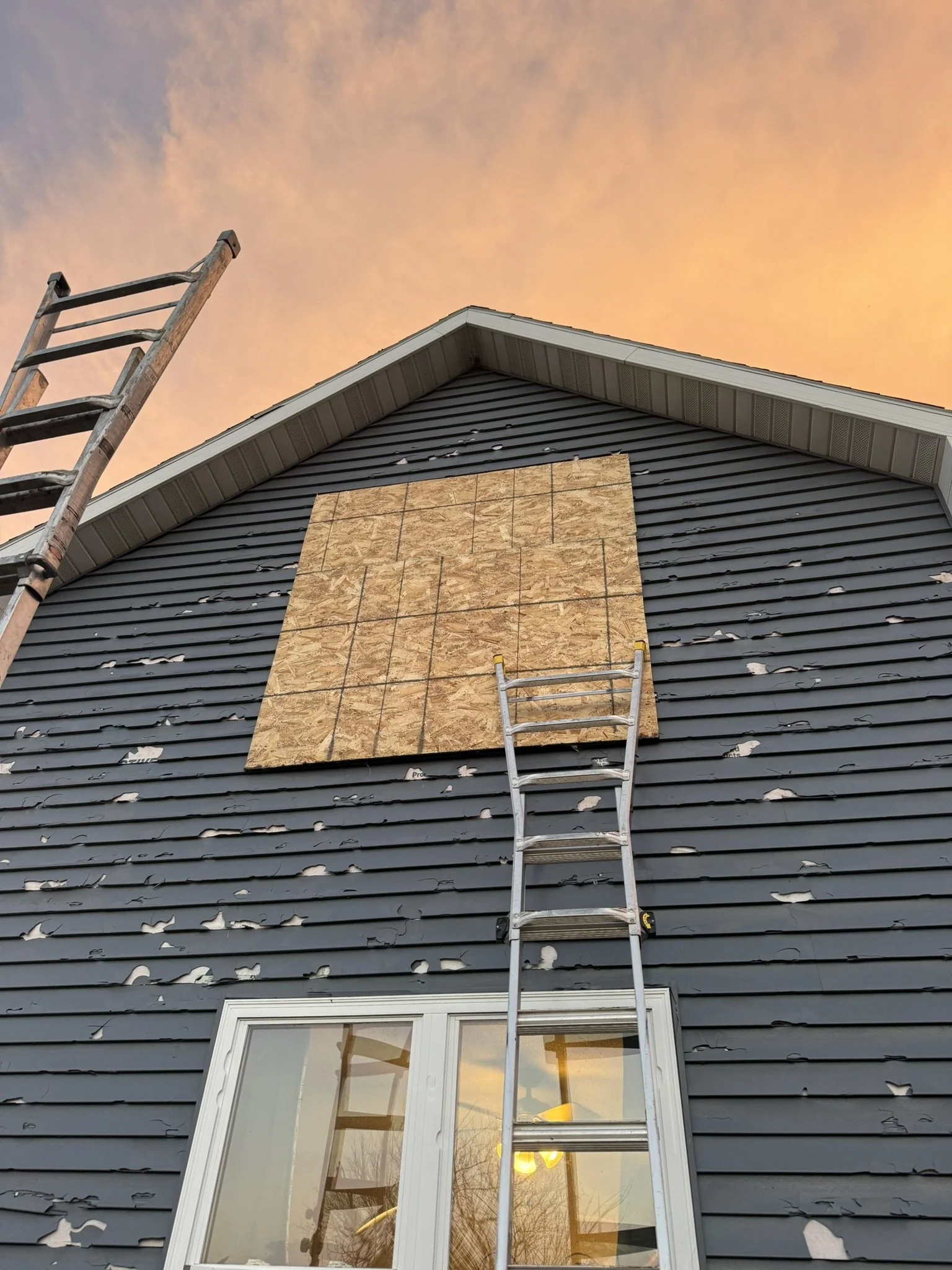 The side of a house with blue siding, peeling paint, a window at the bottom, and a boarded-up section in the middle. There is a ladder leaning against the wall and another ladder nearby. The sky has a warm orange hue with clouds.