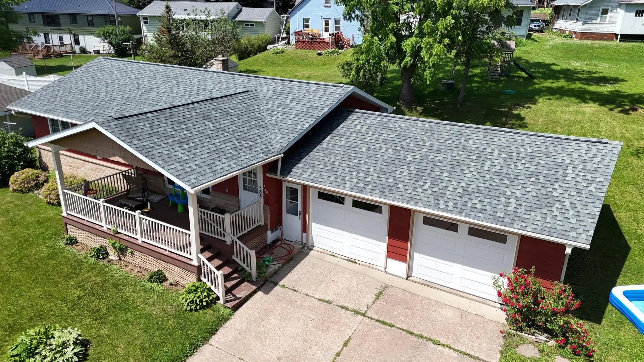 Aerial view of a house with a gray shingle roof, a front porch, and a two-car garage in a neighborhood with other houses, green lawns, and trees.