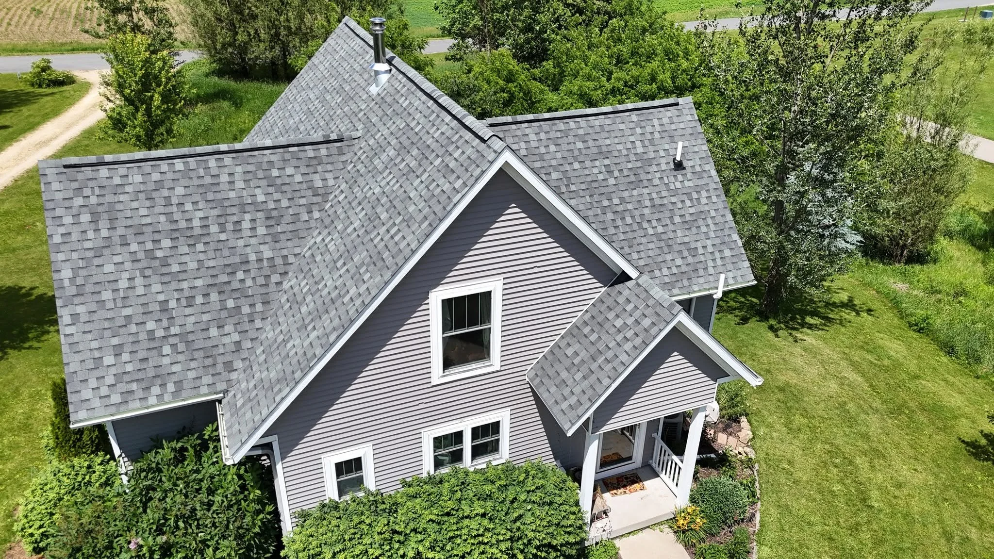 An aerial view of a house with a gray shingle roof, white siding, surrounded by green grass, trees, and a gravel driveway.