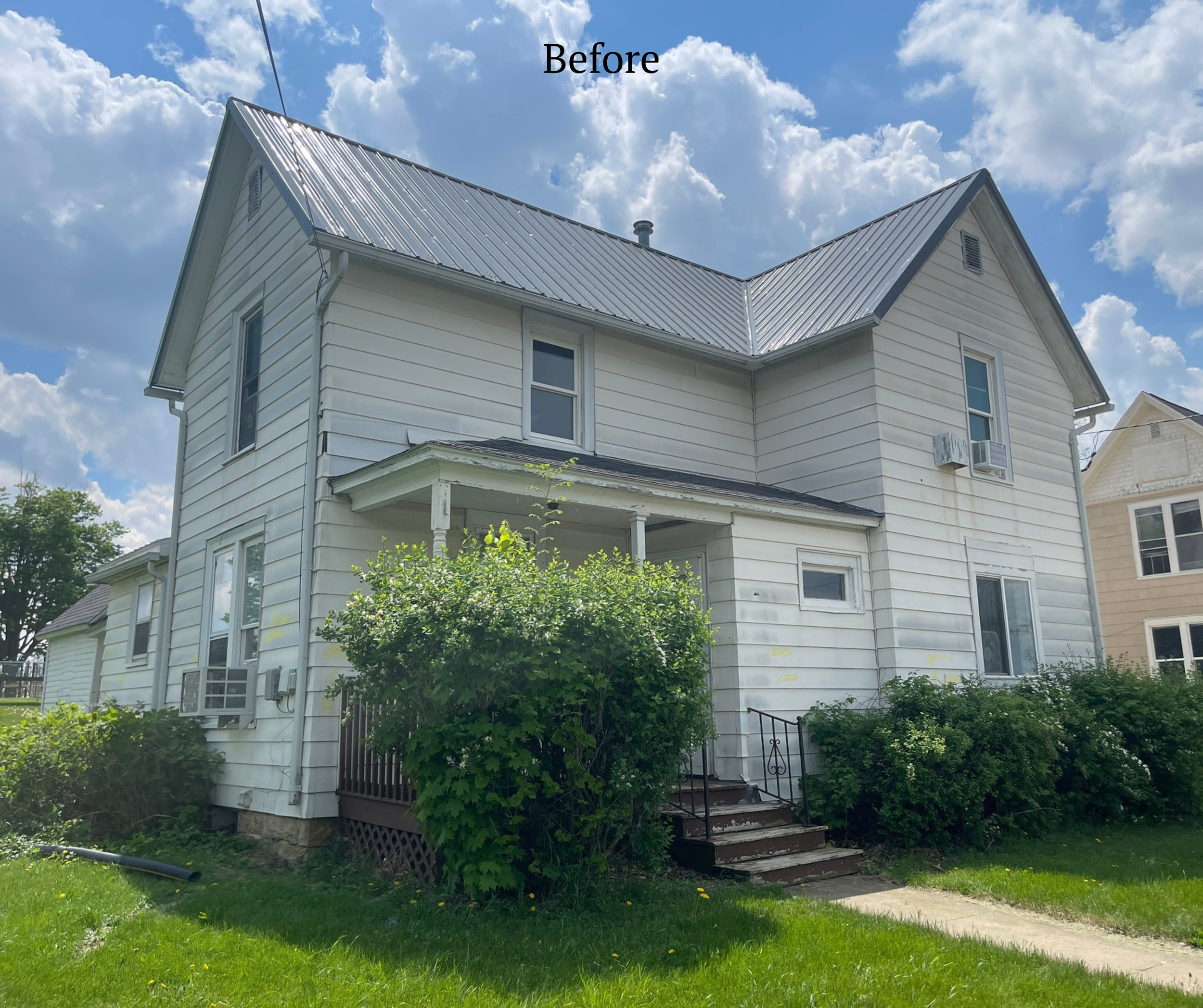 White two-story house with metal roof, surrounded by green bushes and grass, under a partly cloudy sky.