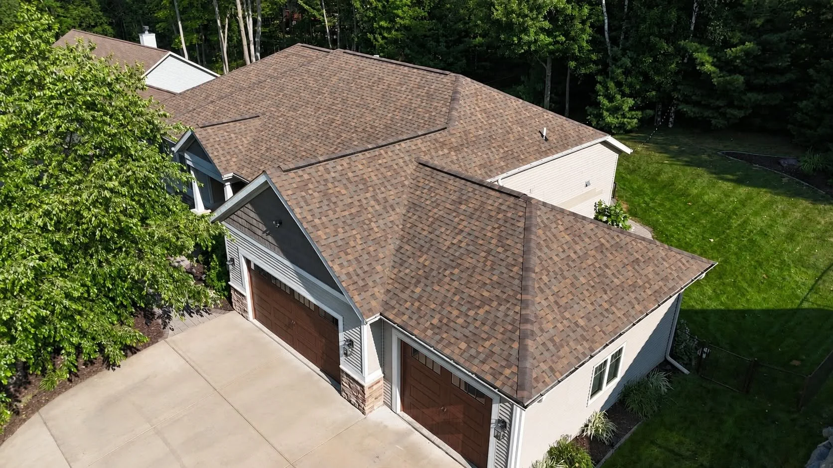 Aerial view of a house with a pitched roof covered in brown shingles, a garage with two doors, and a well-maintained lawn surrounded by trees.