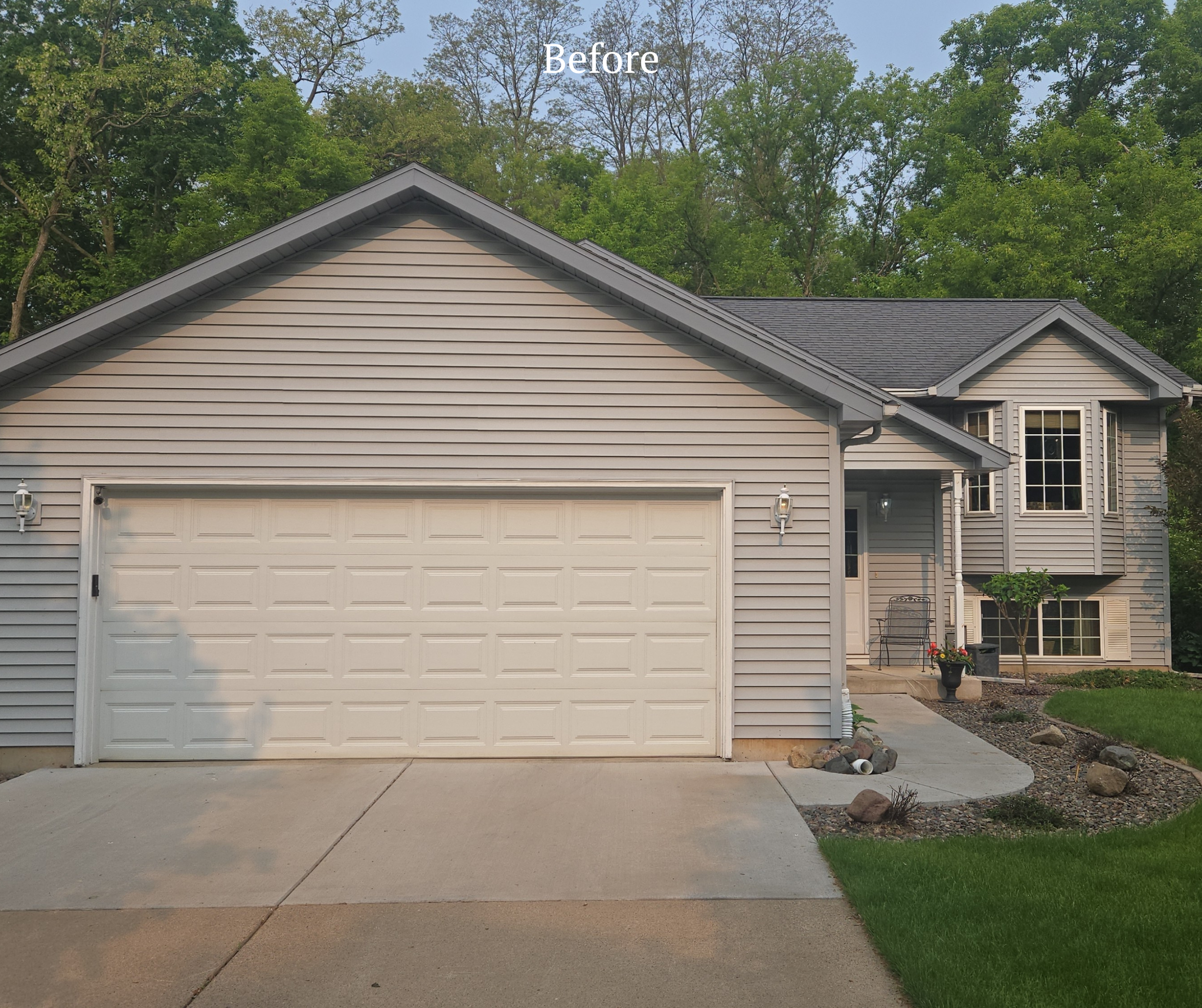 Front view of a house with gray siding, a white garage door, and a small front porch with potted plants, surrounded by a green lawn and trees in the background.