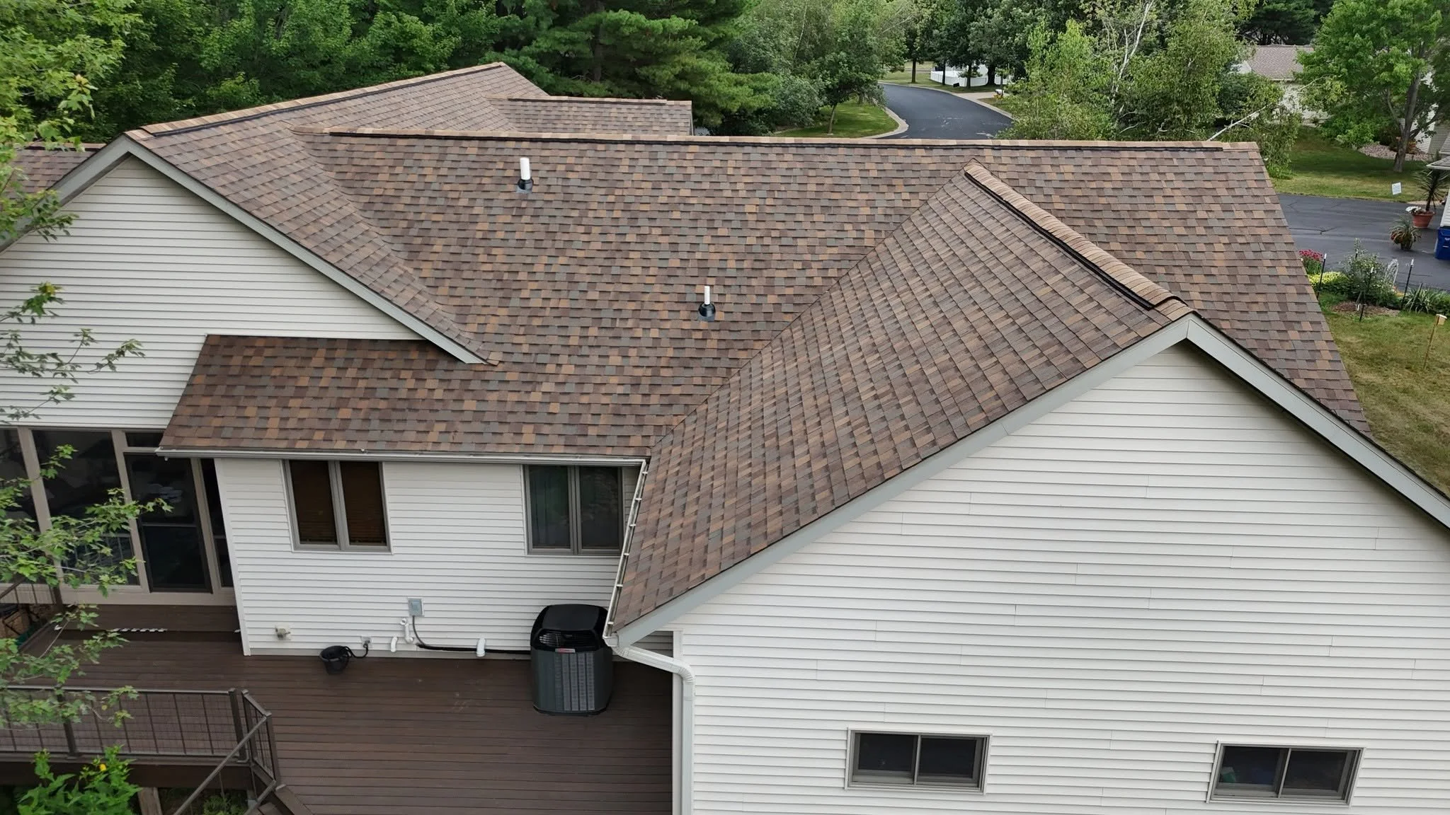 Aerial view of a house with a brown shingled roof and white siding, surrounded by trees and a driveway.