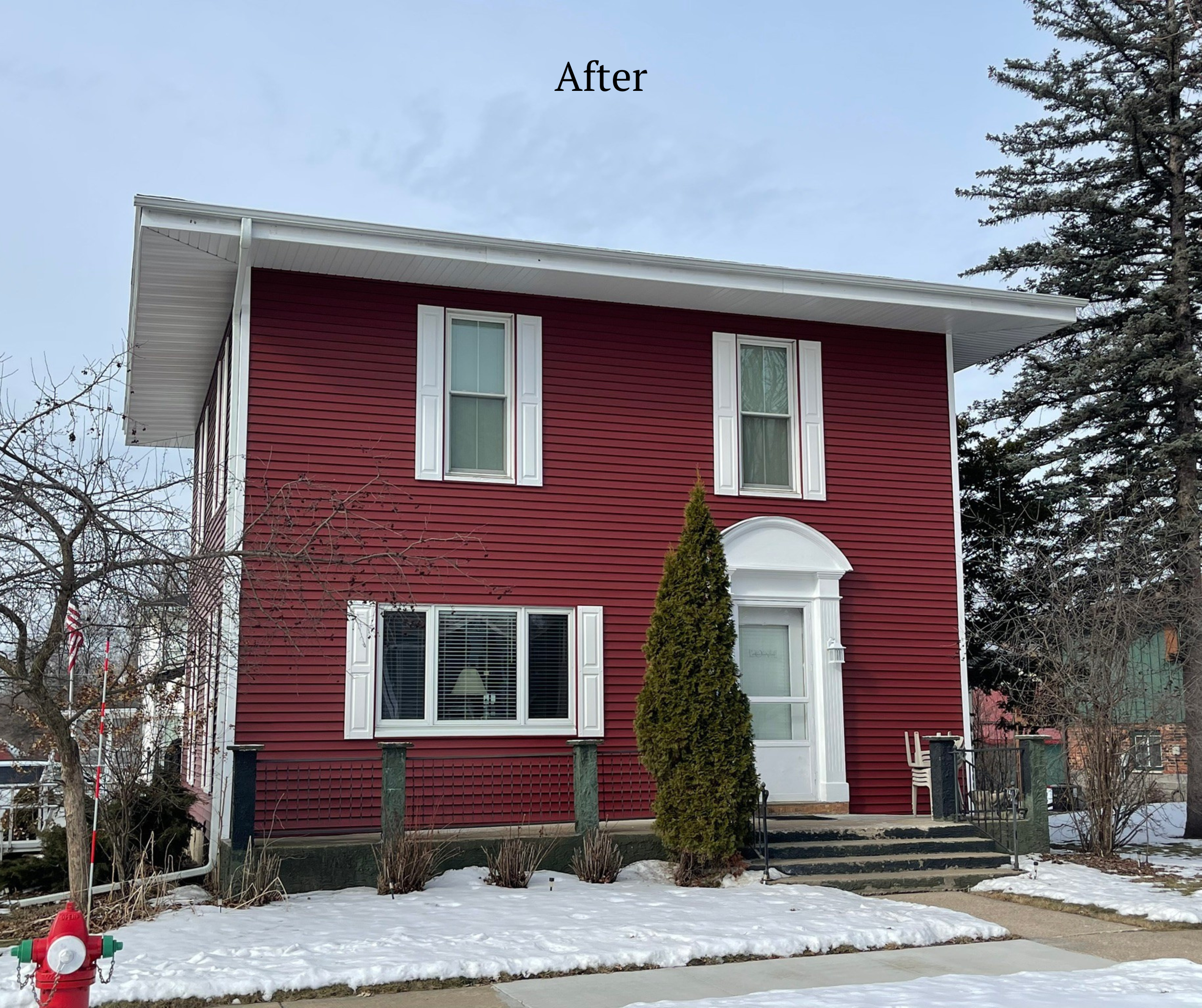 A two-story house with red siding and white trim, featuring four windows and a front door with steps, situated in a snowy landscape with trees around.