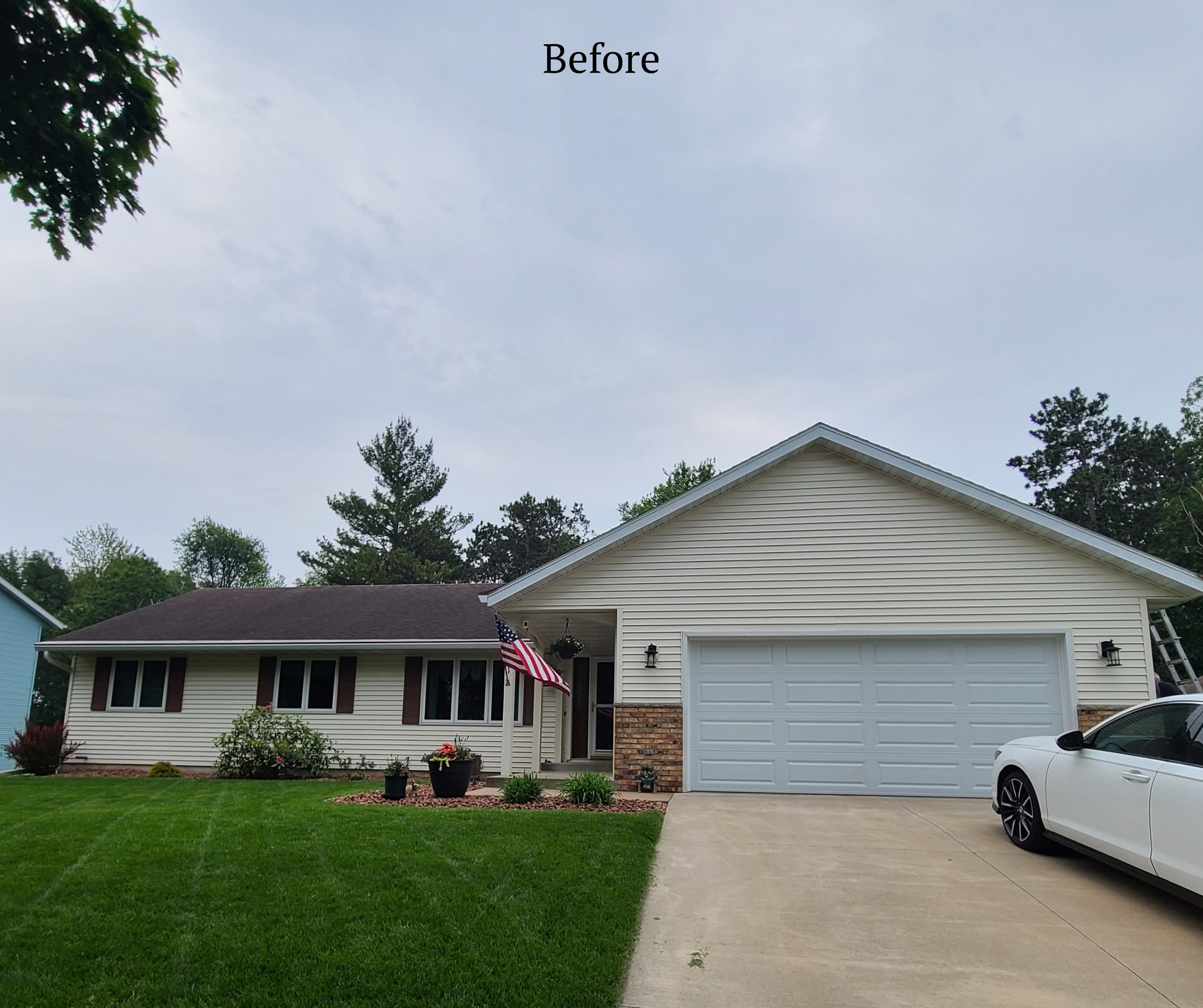 A house with beige siding and a two-car garage, surrounded by a well-maintained lawn with potted plants and flowers, a flag hanging near the front door, and a white car parked in the driveway under an overcast sky.