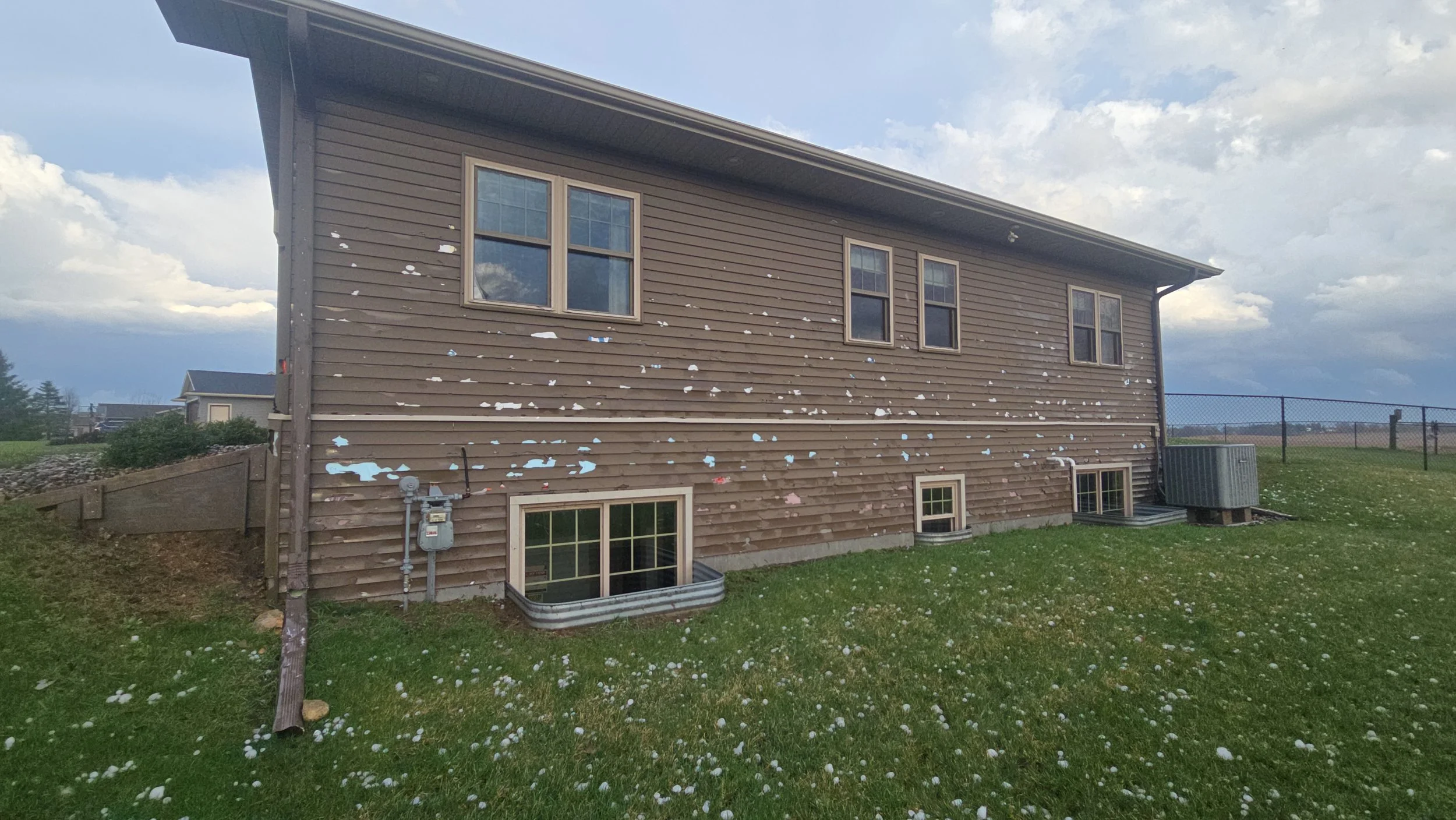 Back of a two-story house with brown siding, showing peeling paint, windows, and exterior utilities in a grassy yard under a partly cloudy sky.