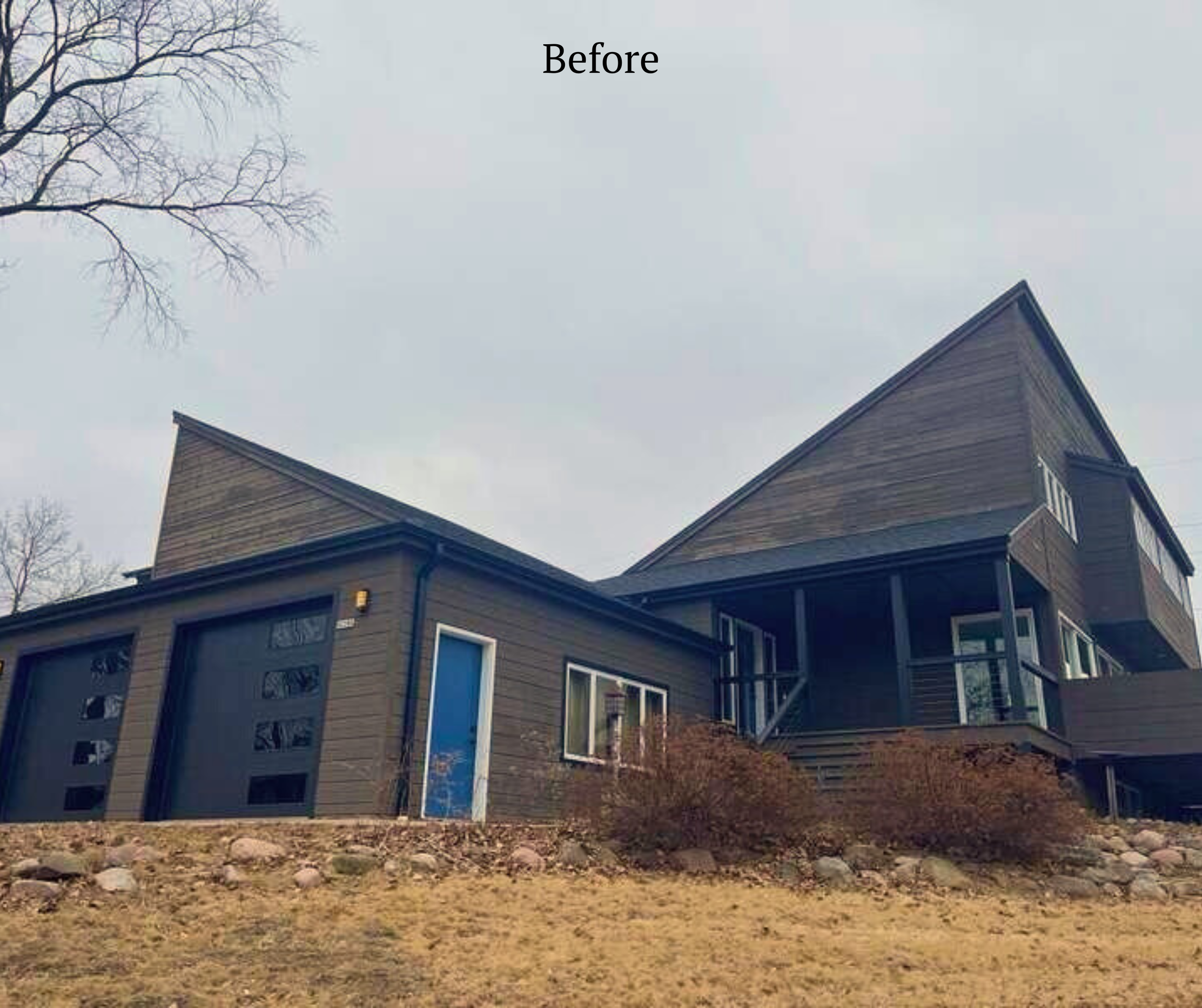A modern house with dark gray exterior, multiple levels, large windows, and a sloped roof, situated on a rocky hill with leafless trees in the background and an overcast sky.