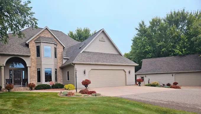 Aerial view of a modern suburban house with a gray shingled roof, white siding, a double garage, and well-maintained landscaping, situated in a green rural area.