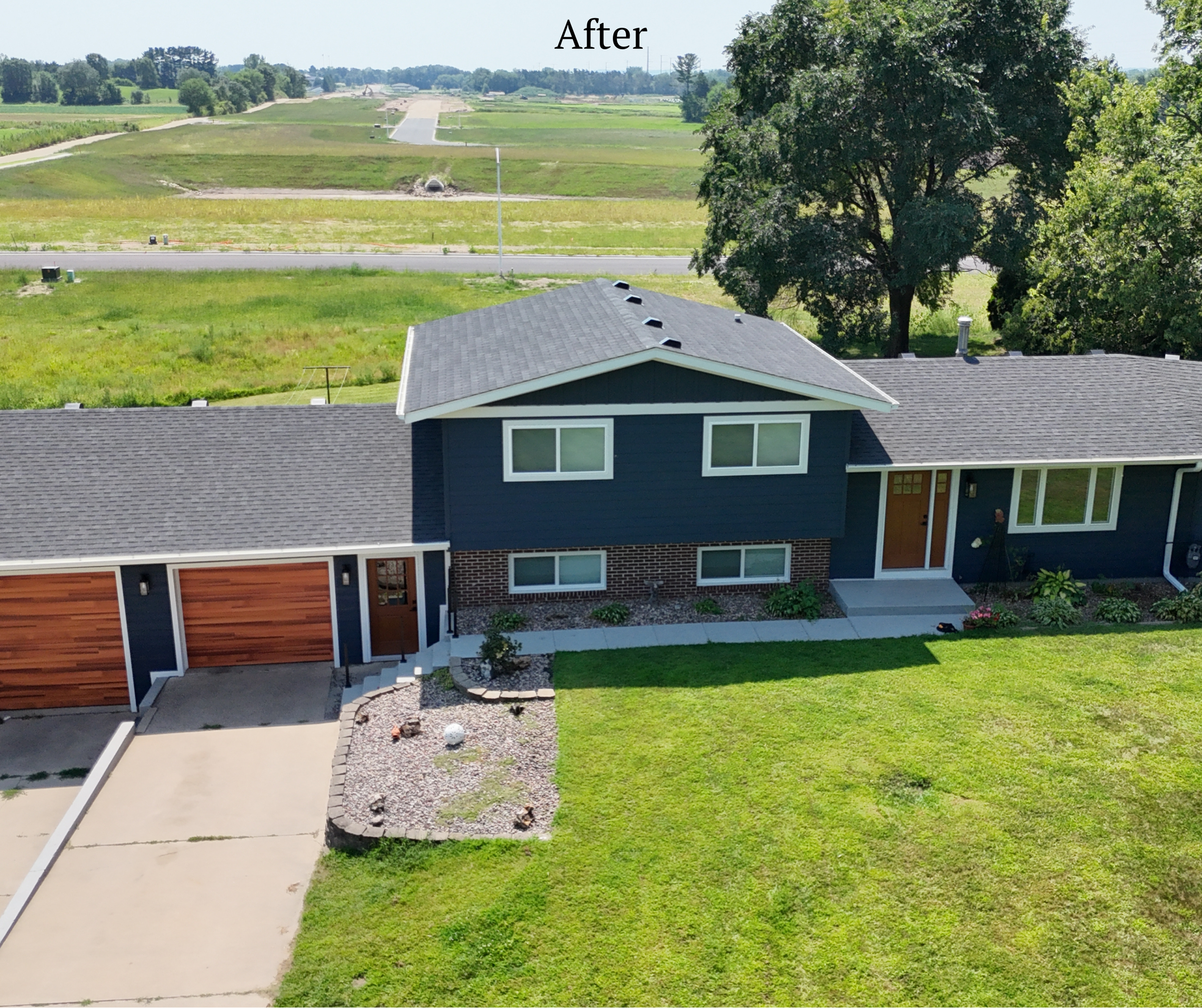 A modern two-story house with dark blue exterior, white window frames, a gray roof, and a front porch with a wooden door. The front yard has green grass, a small landscaped area with rocks and plants, and a concrete driveway. Trees are visible in the