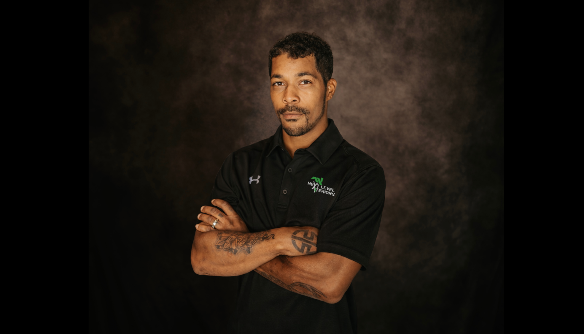 A man with short black hair and a goatee standing against a dark, textured backdrop. He is wearing a black polo shirt with a logo on the left chest that reads 'Next Level Interiors' and has tattoos on his left forearm. He is smiling slightly and looking at the camera.