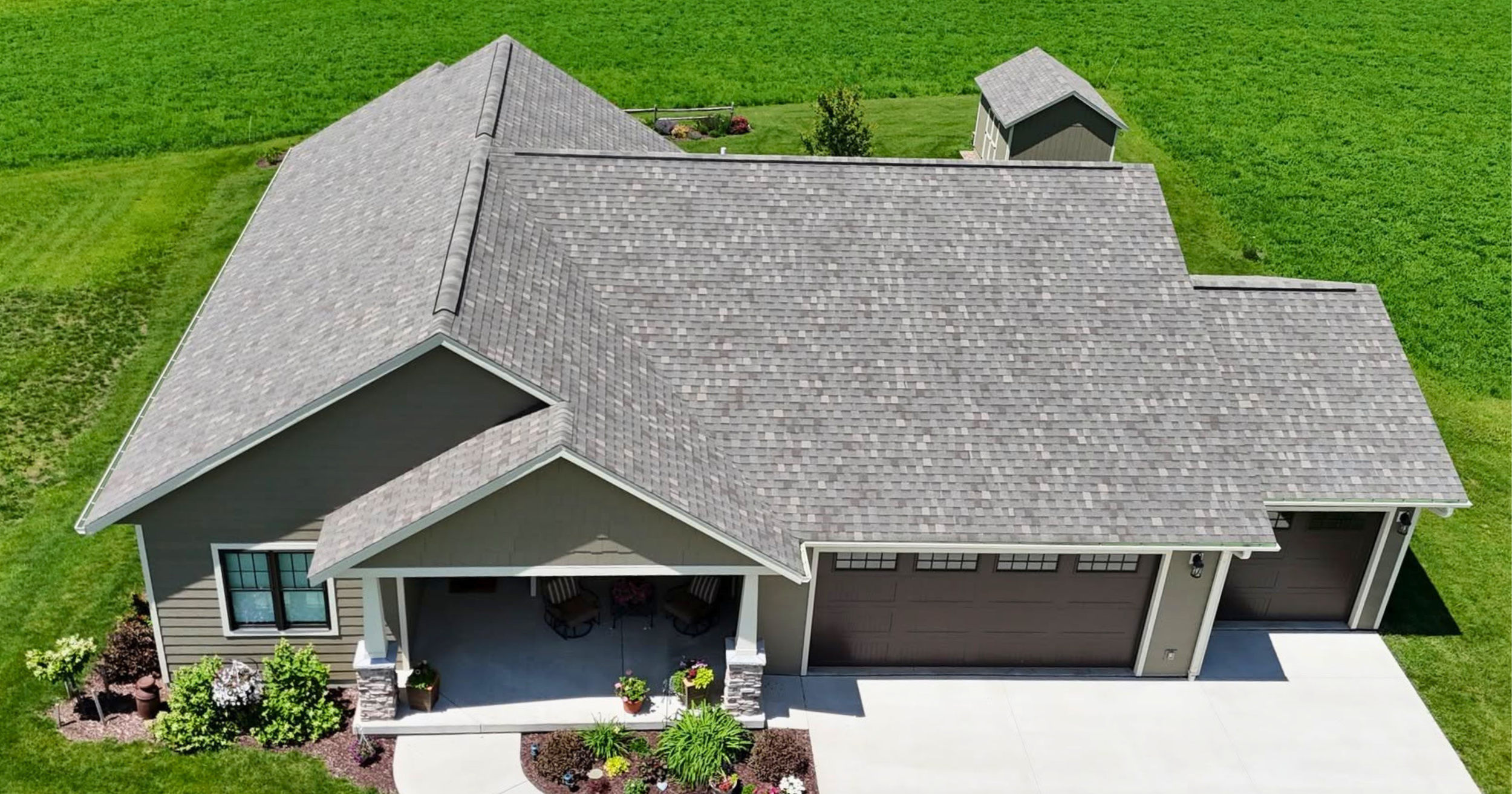 An aerial view of a house with a gray shingle roof, beige siding, a front porch with white columns, surrounded by a manicured lawn and landscaped garden.
