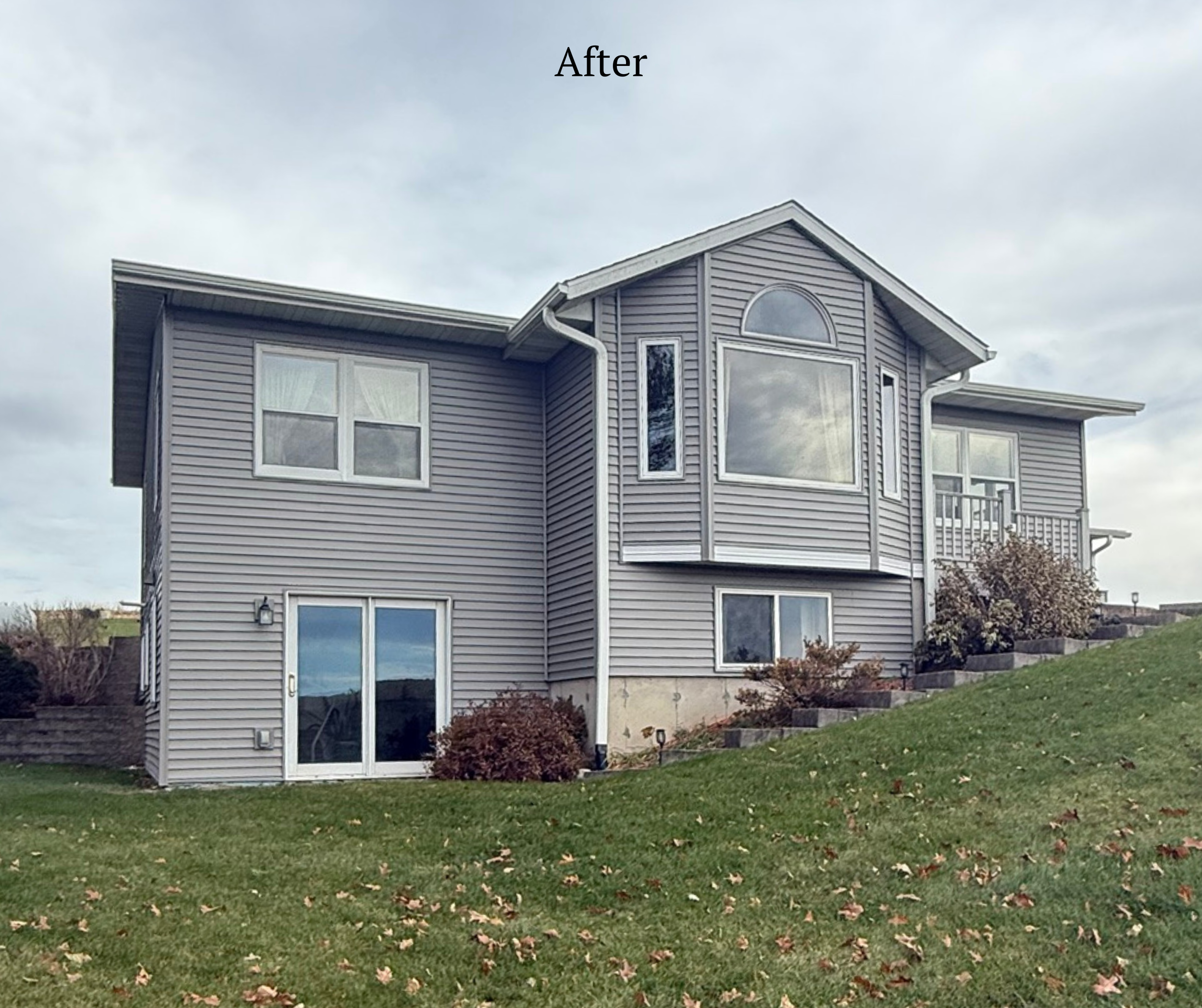The image shows the back of a two-story house with gray siding, large windows, a small balcony, and landscaped bushes on a grassy slope under a cloudy sky.