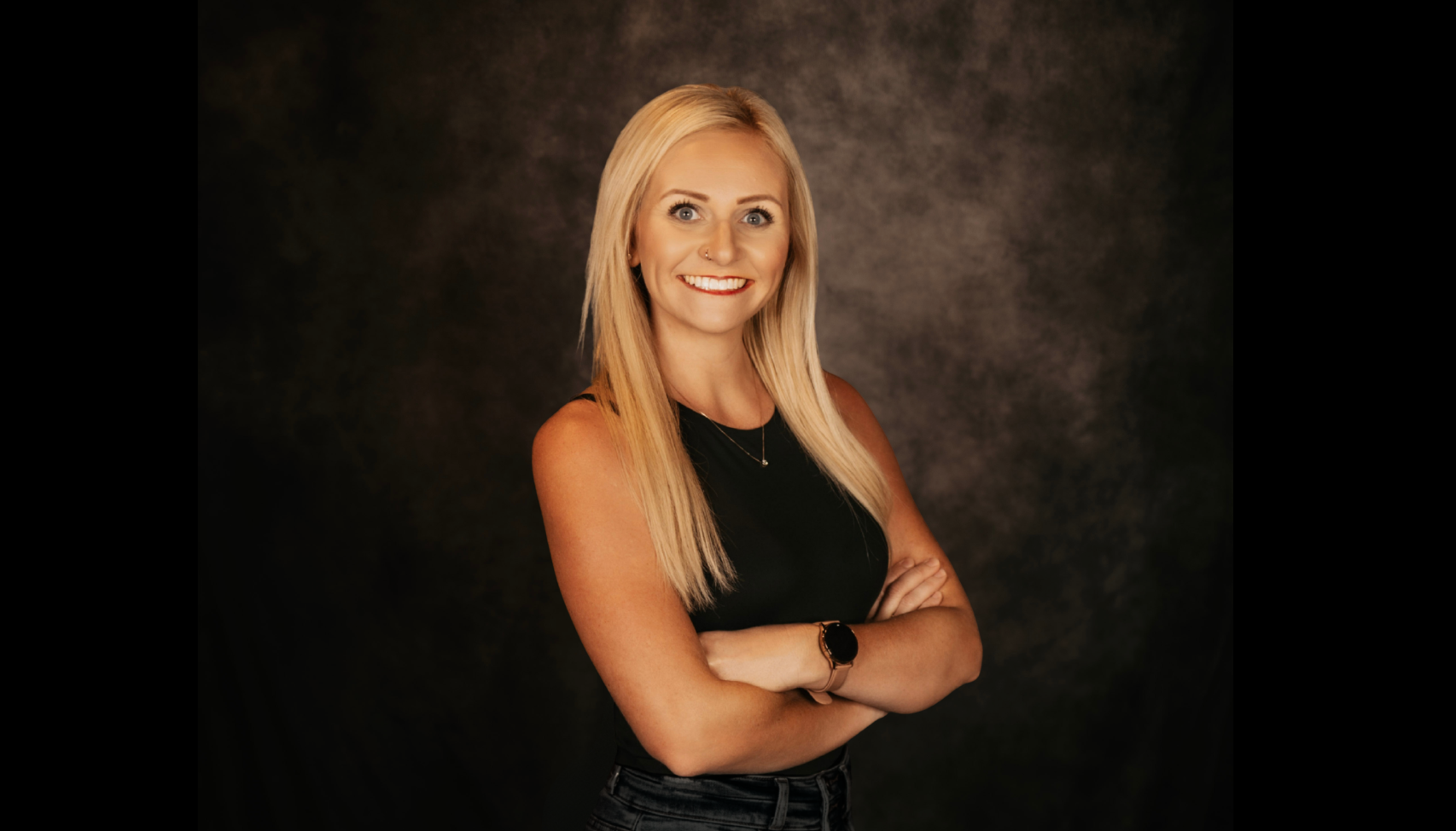 A young woman with long blonde hair, blue eyes, wearing red lipstick, a small nose piercing, and earrings. She is dressed in a black sleeveless top and blue jeans, standing against a dark background.