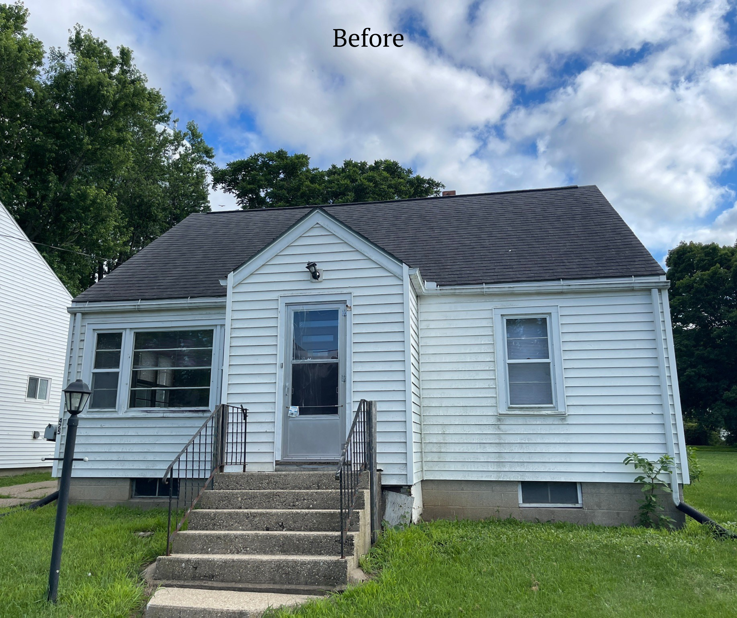 A small white house with a black shingled roof, a front staircase with black handrails, a screen door, two windows on the front, and a grass lawn with a lamp post. The sky is partly cloudy with blue patches.