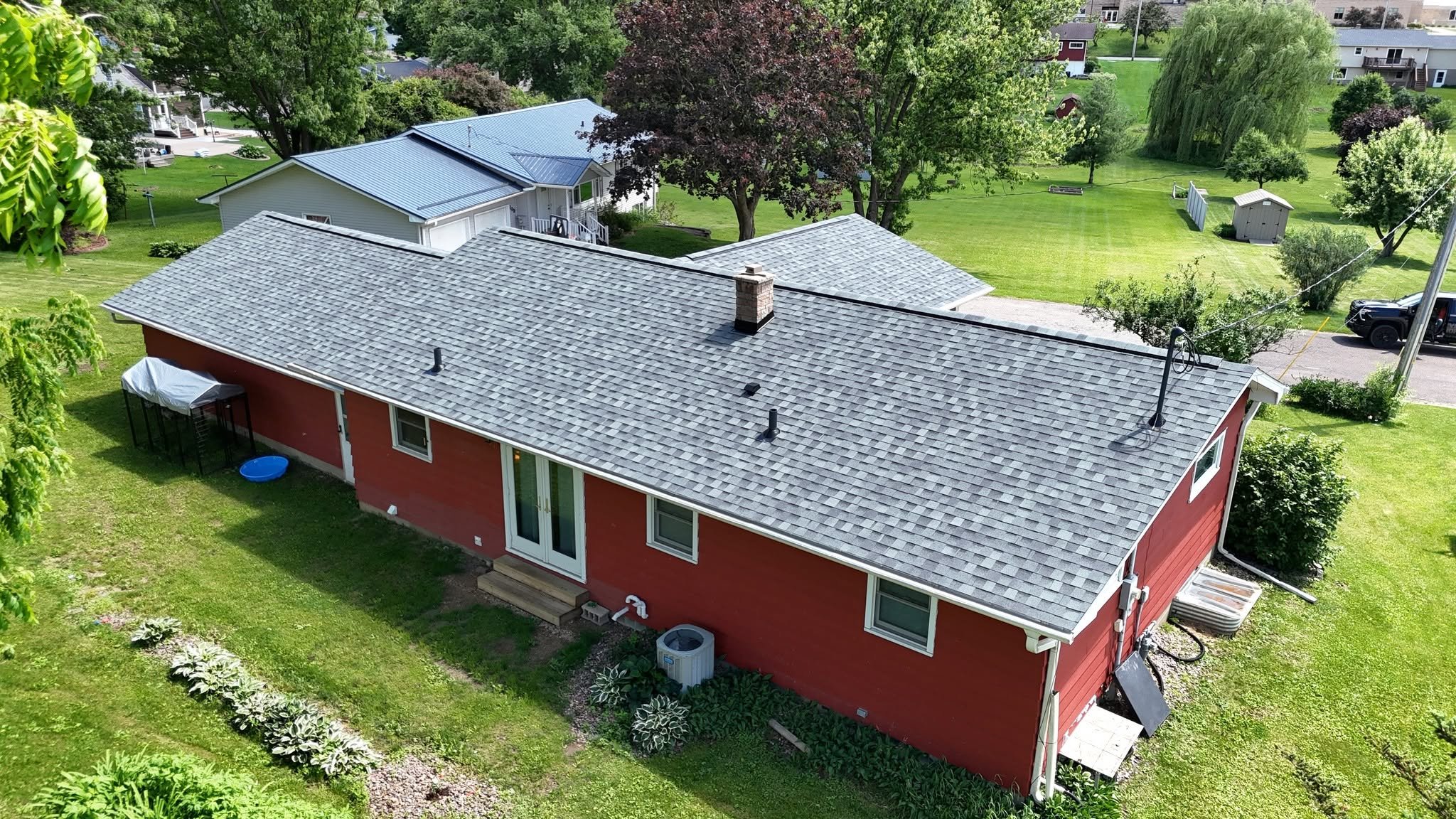 Aerial view of a red house with a gray shingled roof, surrounded by green lawns, trees, and neighboring houses in the background.