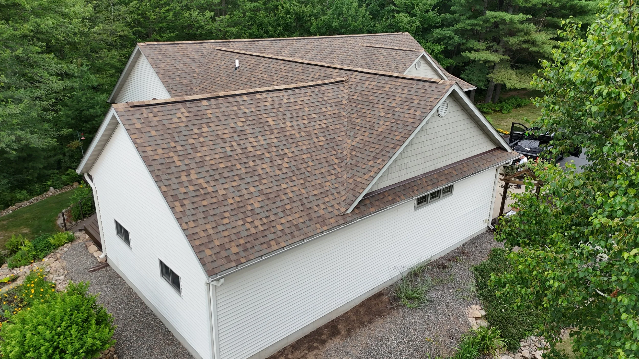 Aerial view of a house with a brown shingle roof, white siding, surrounded by greenery and trees.
