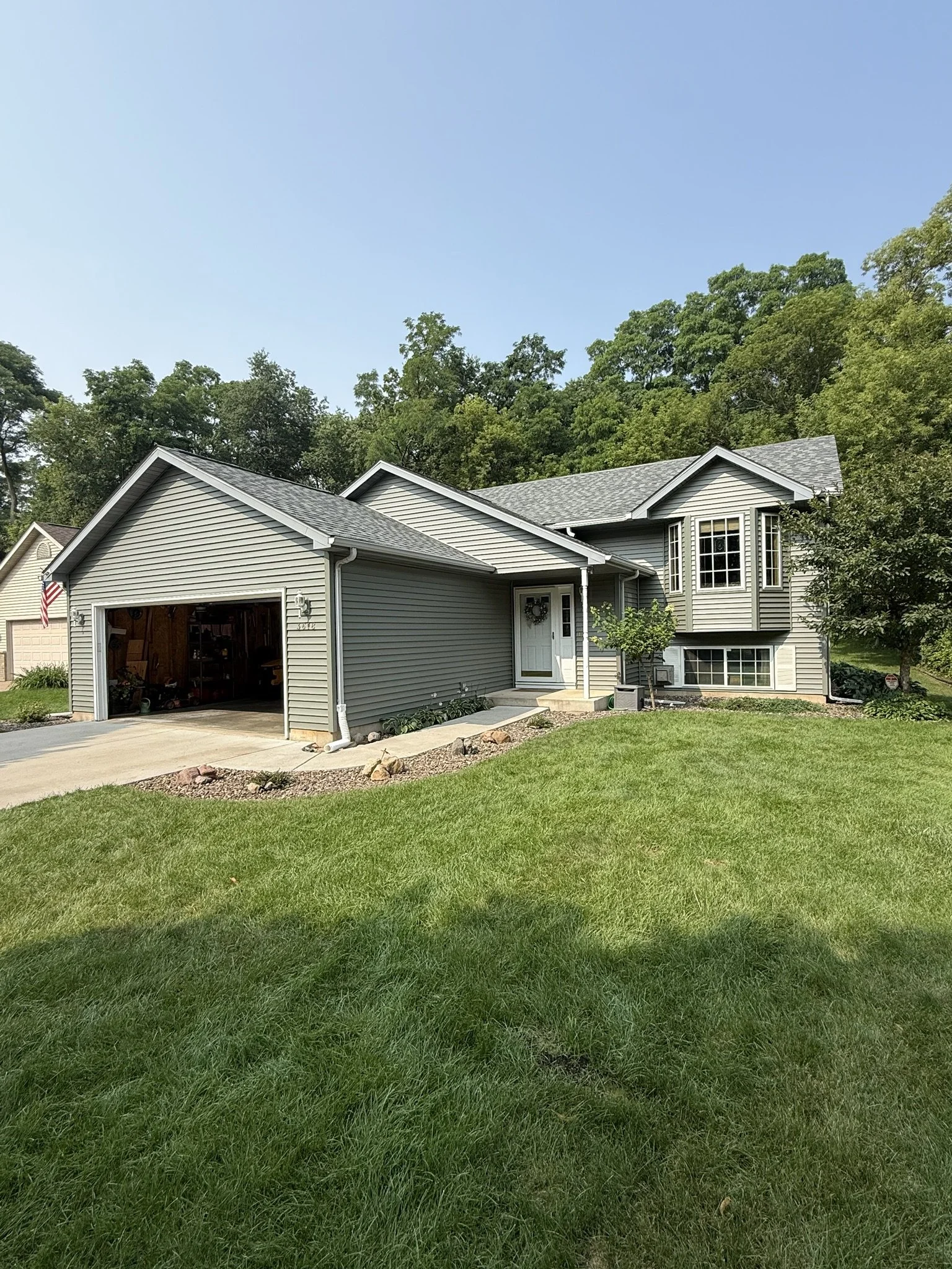 Front view of a gray suburban house with a two-car garage, white door, and well-maintained lawn, surrounded by green trees.