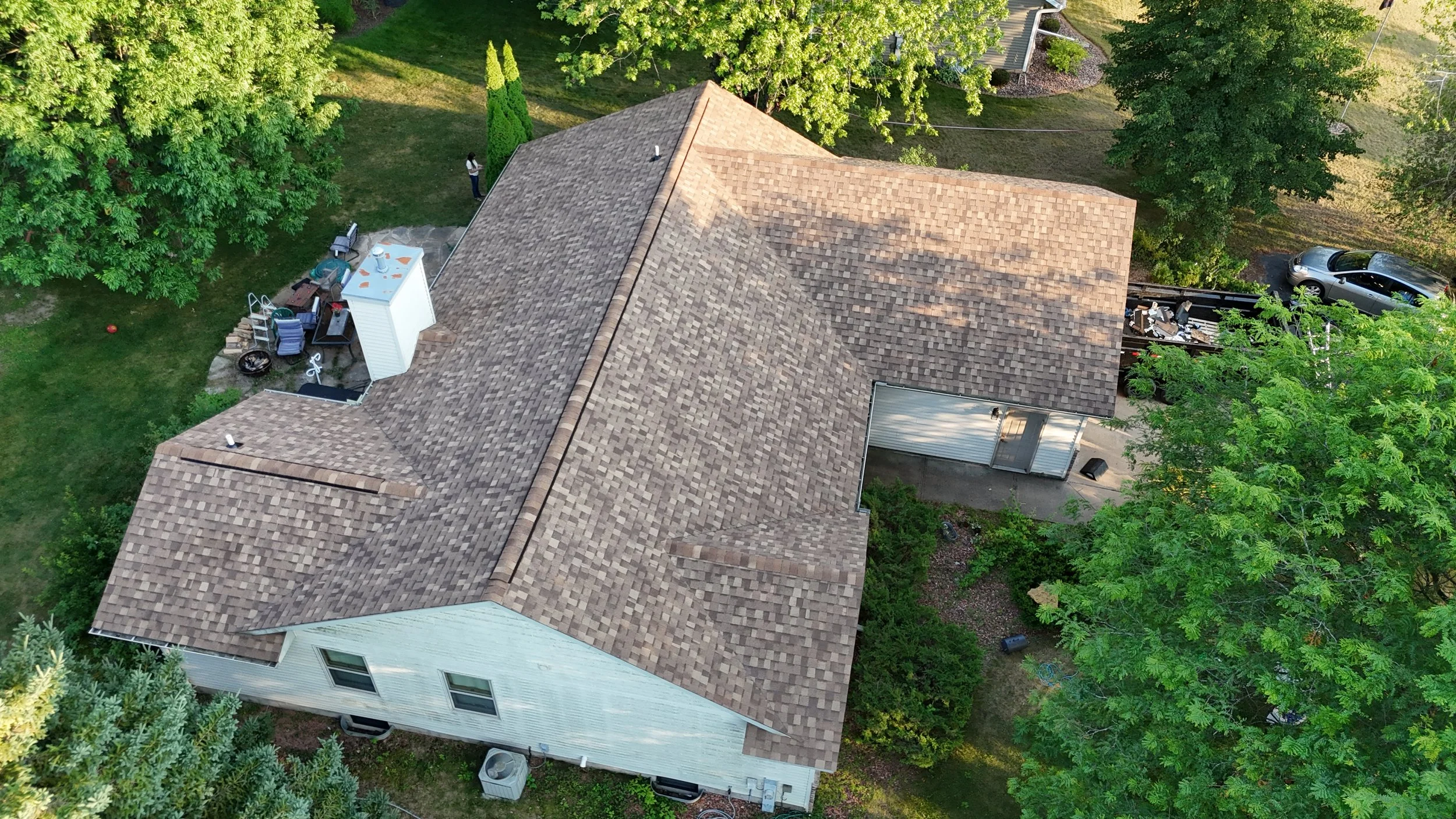 An aerial view of a house with a brown shingle roof, surrounded by green trees and a yard with scattered outdoor items.
