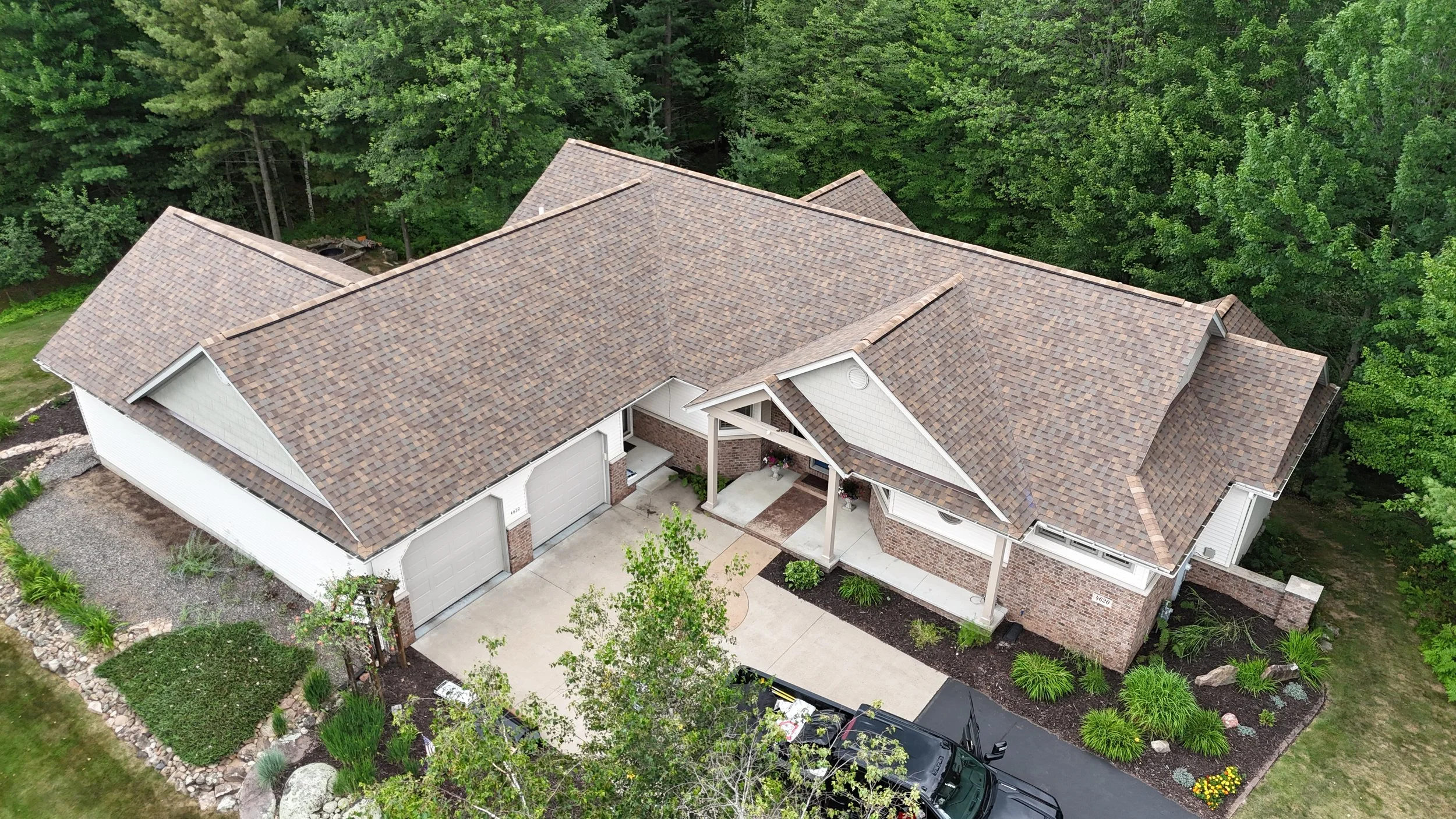An aerial view of a house with a large, multi-gabled roof covered with brown shingles, surrounded by green trees and landscaped yard.
