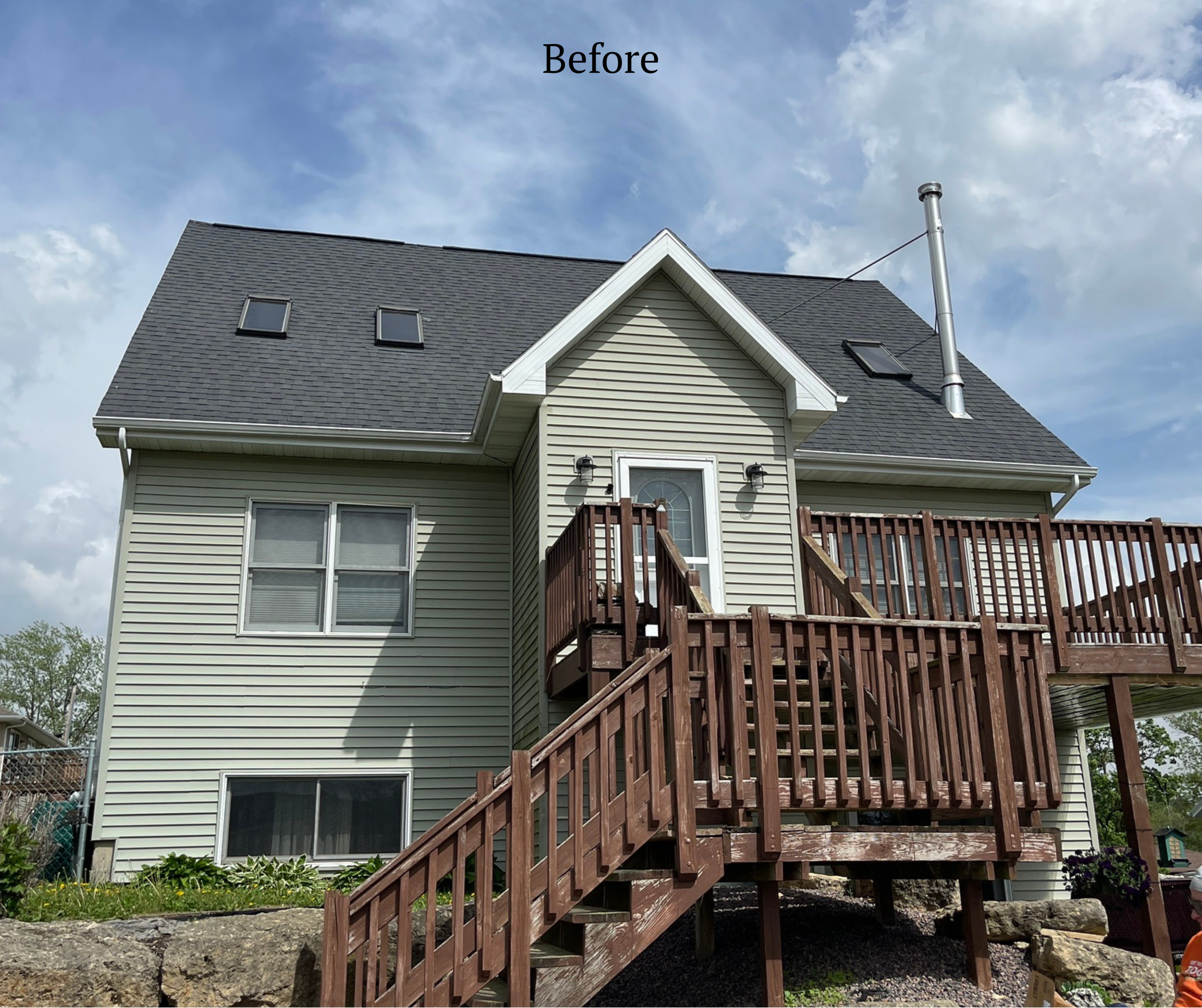 The image shows the back of a two-story house with beige siding and a dark gray roof. There is a new wooden deck and staircase leading to a door on the second floor, with two outdoor wall lights beside the door. The house has two skylights on the roo