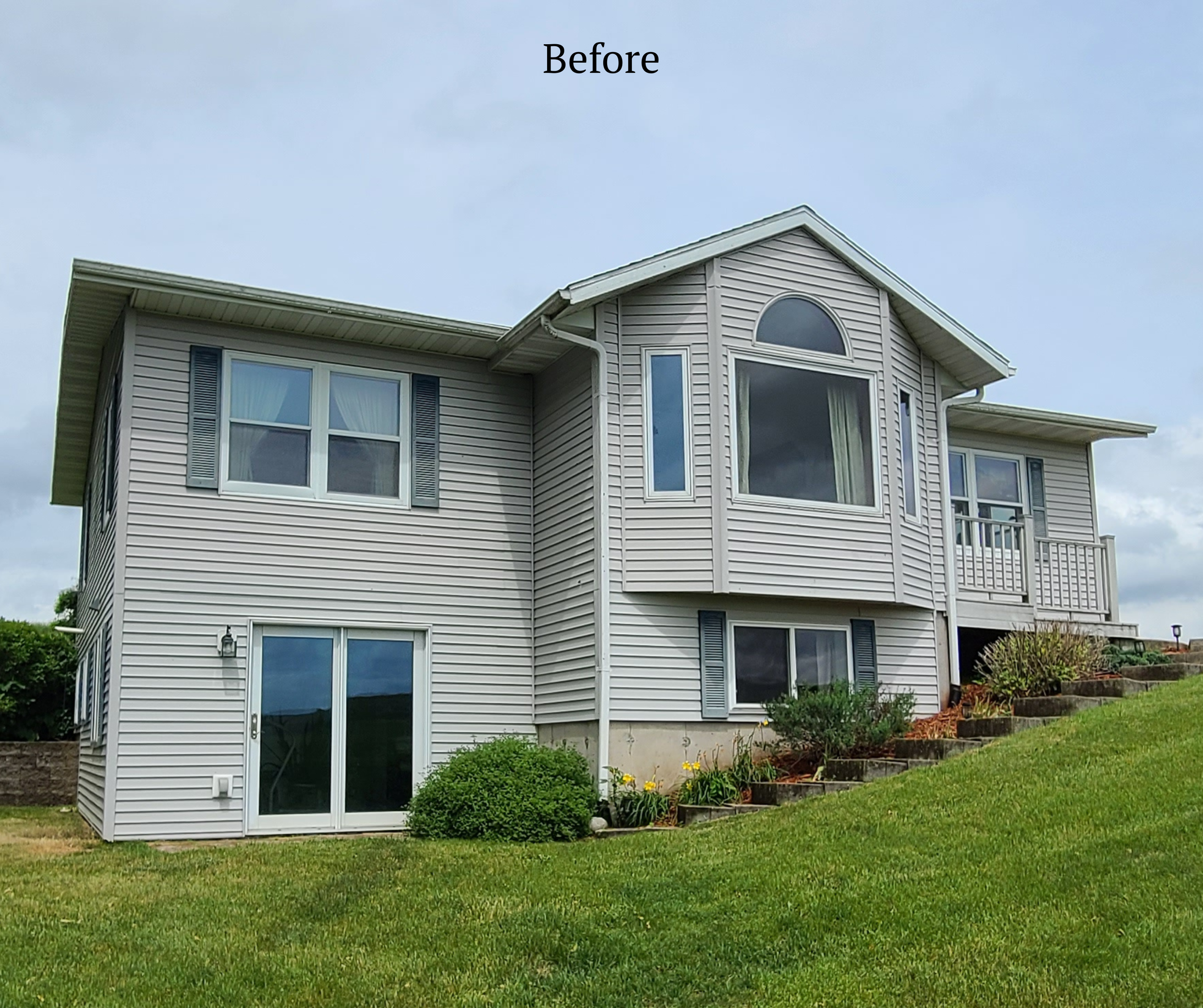 A two-story house with beige vinyl siding and dark blue shutters, situated on a grassy lawn with landscaped bushes and stairs leading to a side porch.