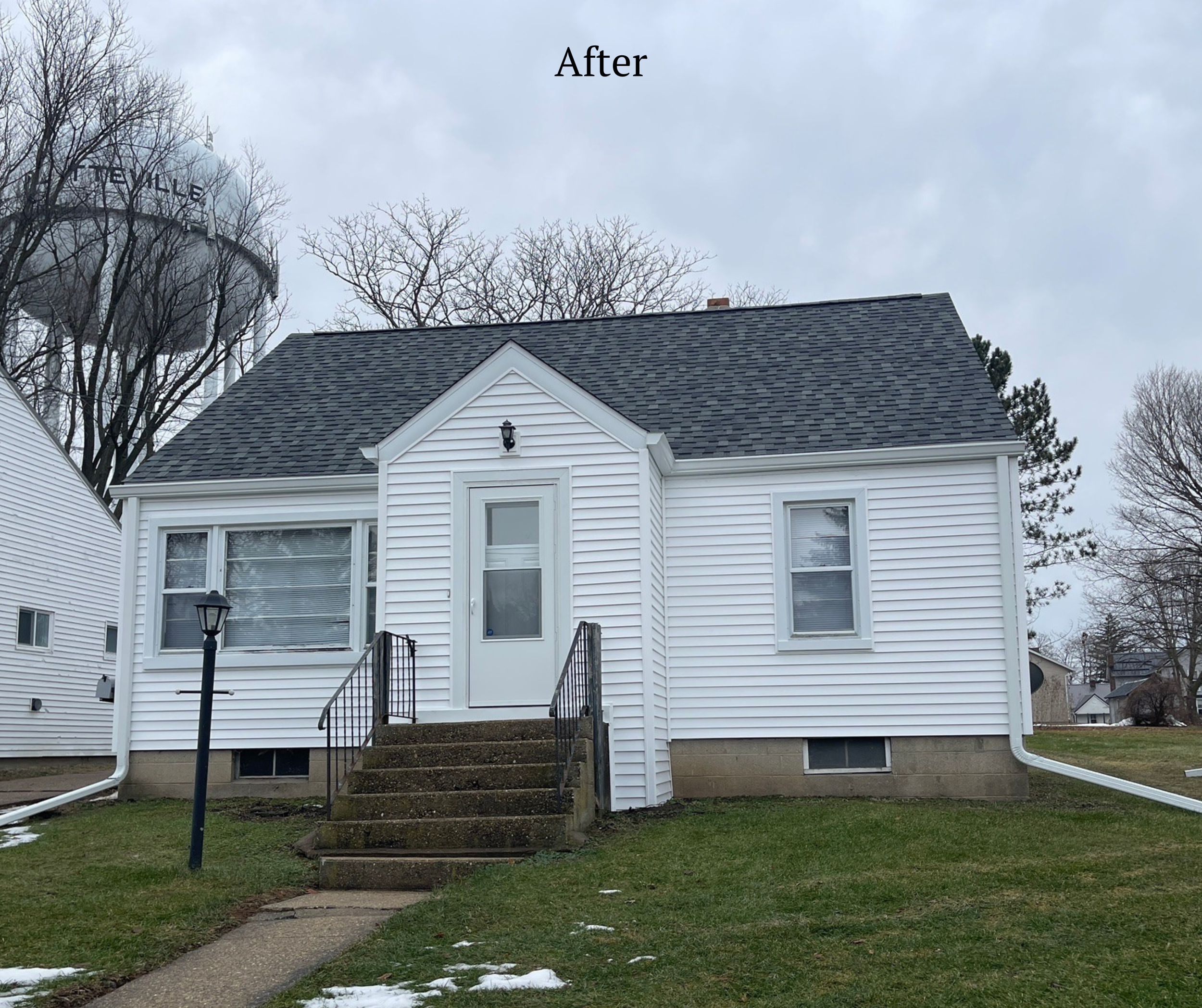 A small white house with a front steps and a door, and four visible windows. There is a lamp post on the lawn near the stairs and an overcast sky.
