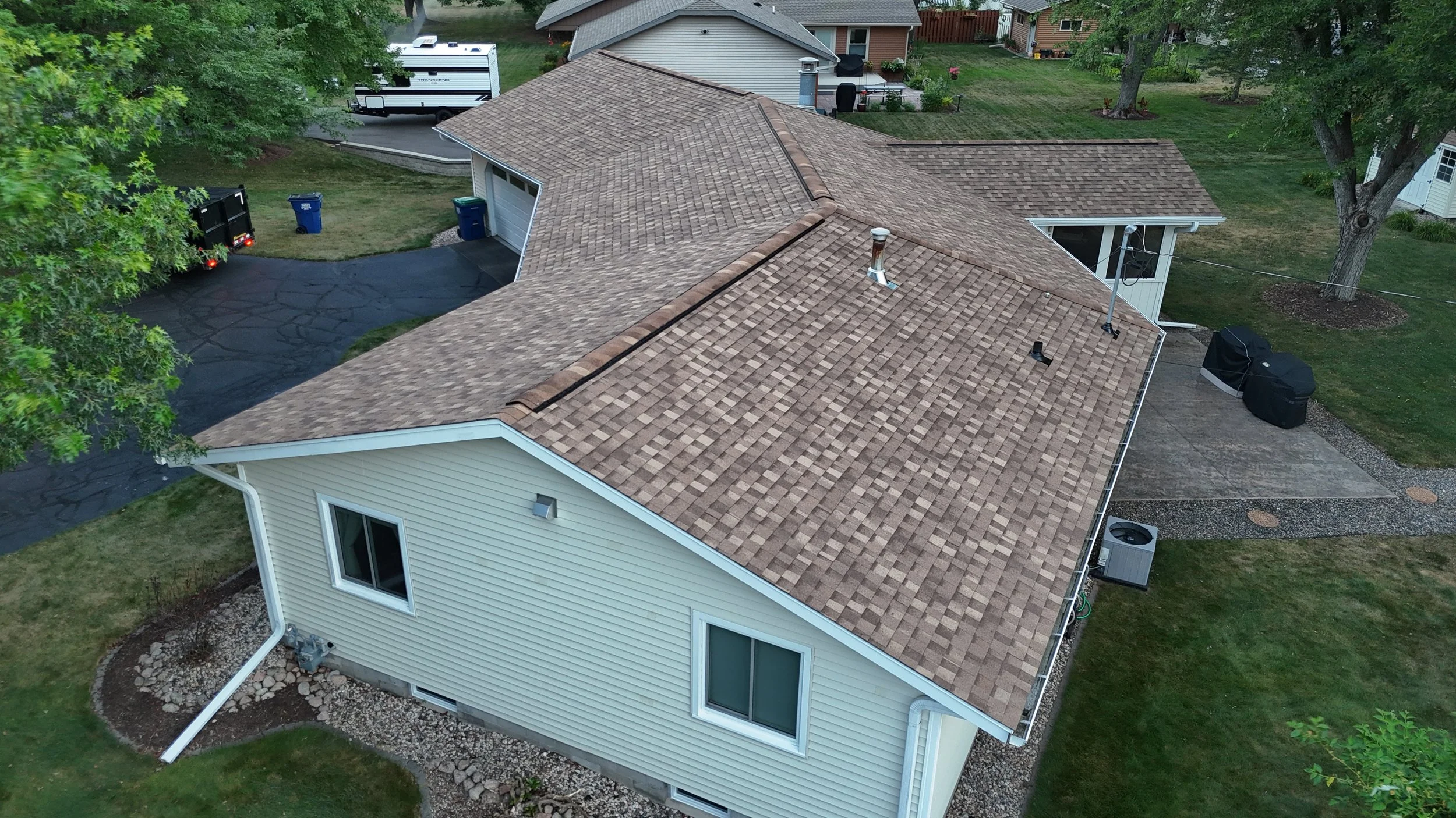 Aerial view of a house with a brown shingle roof, neighboring houses, trees, and a driveway with a trailer and trash bins.
