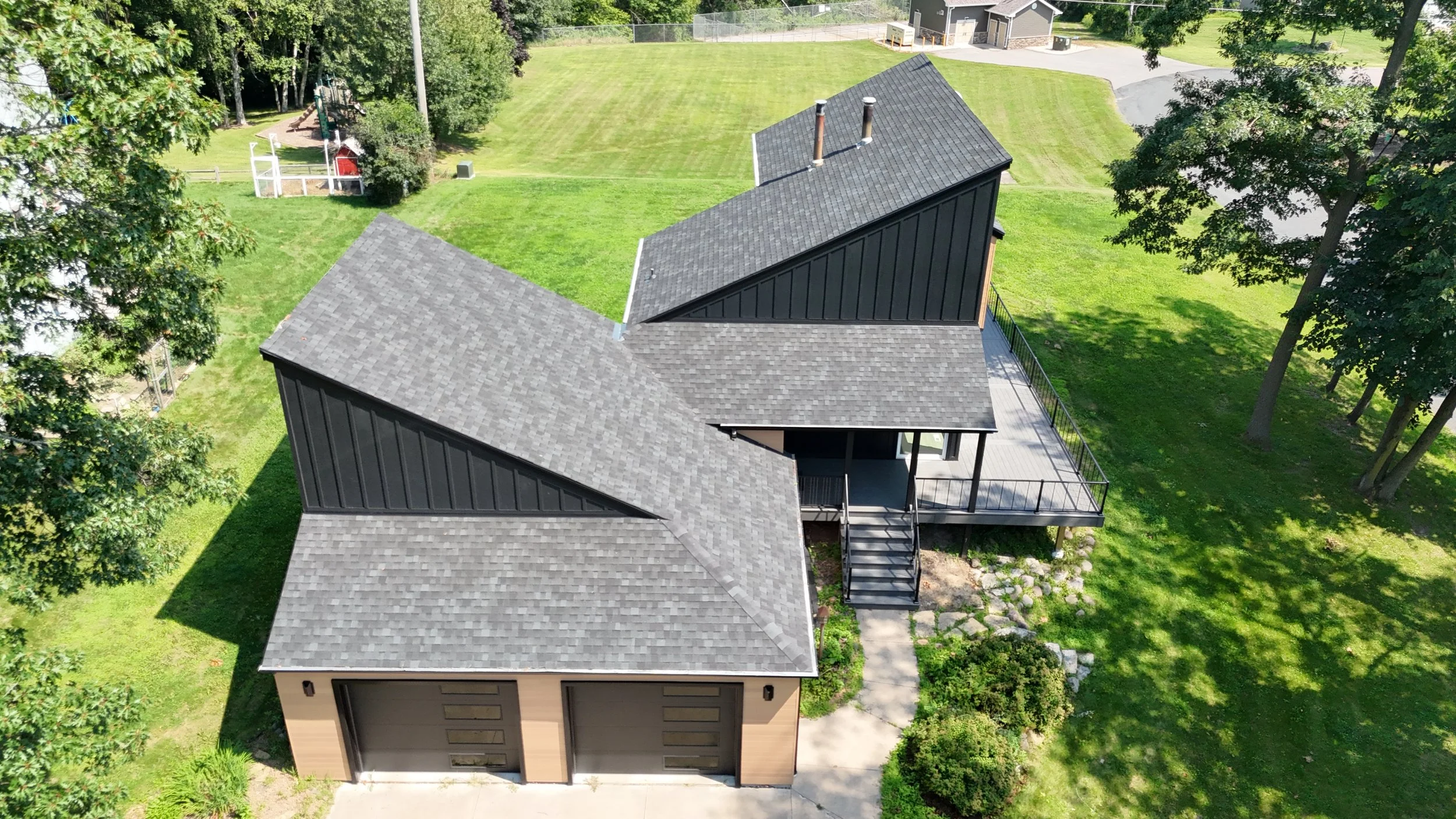 Aerial view of a modern house with a gray shingle roof, black exterior walls, and a balcony. The house is surrounded by green lawn and trees.