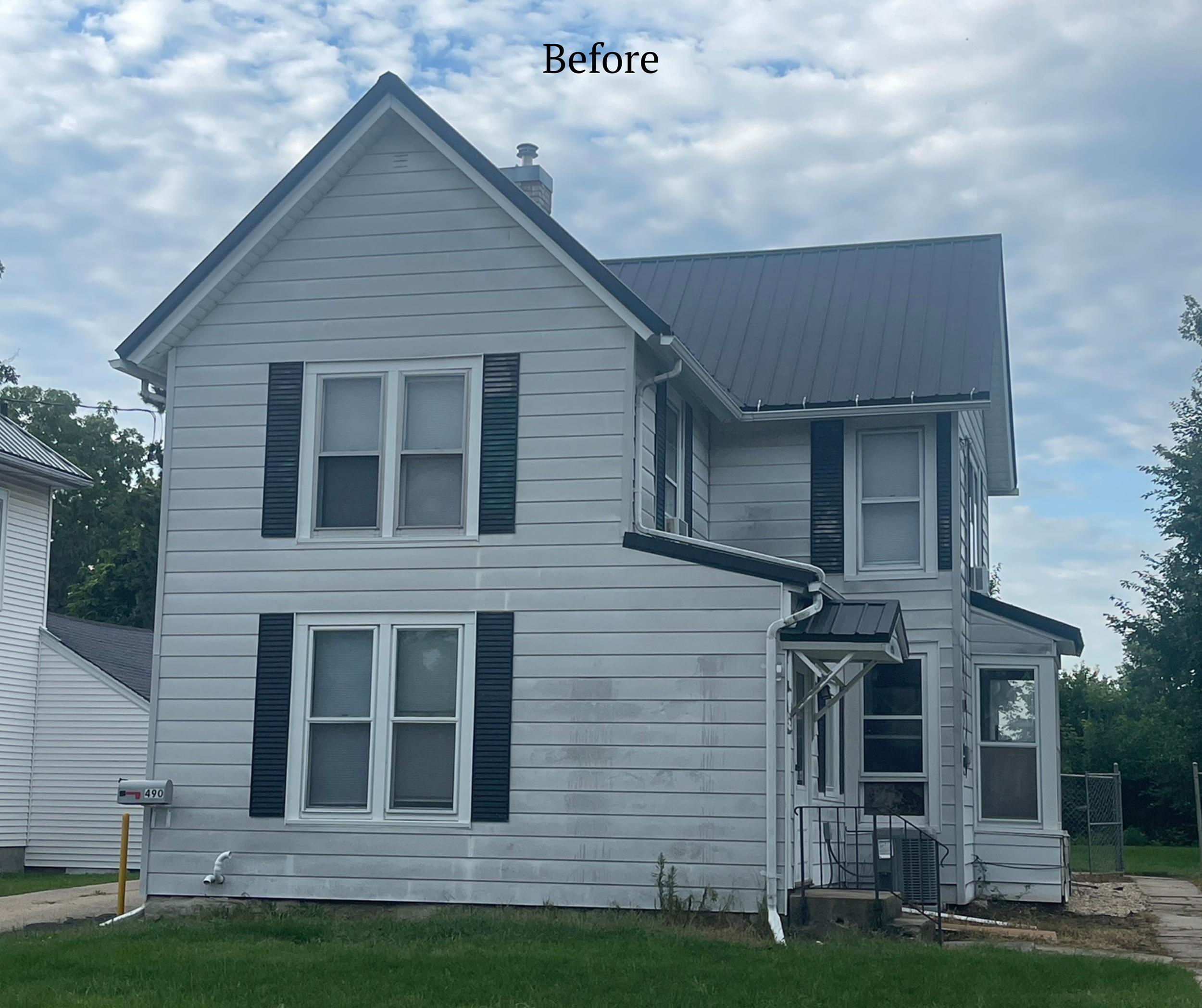 Front view of a white, two-story house with black shutters, a dark metal roof, and a small porch with steps. The house appears slightly dirty or stained on the exterior walls. The sky above is partly cloudy.