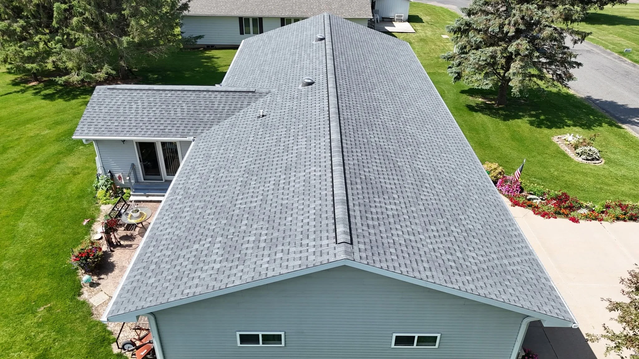 Aerial view of a house with gray shingle roof, green lawn, trees, and a small patio with outdoor furniture.