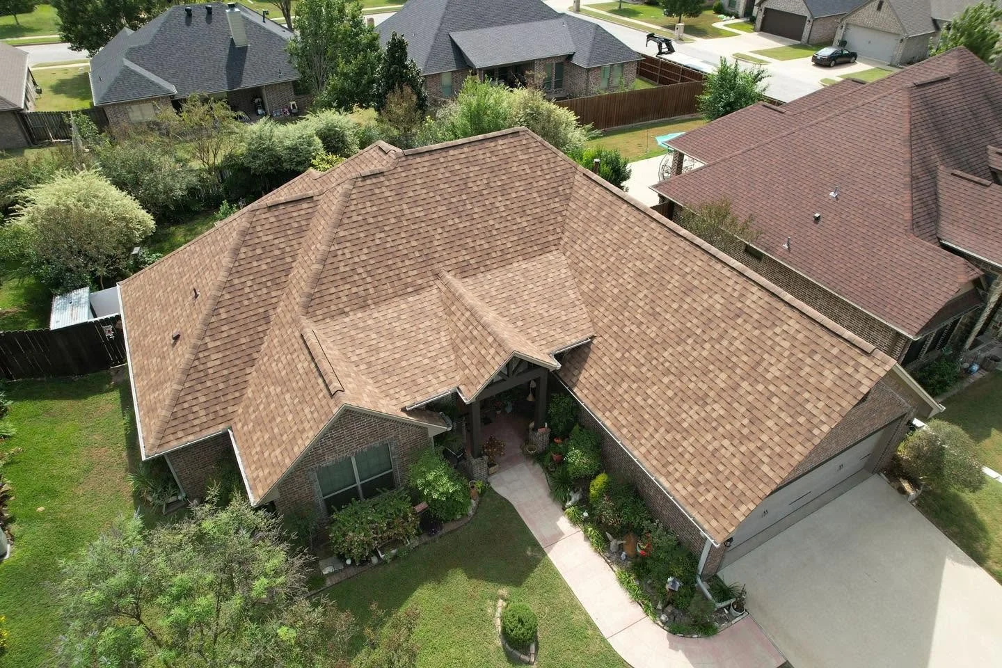 An aerial view of a suburban house with a brown tiled roof, surrounded by green lawns and trees, with neighboring houses and driveways visible.