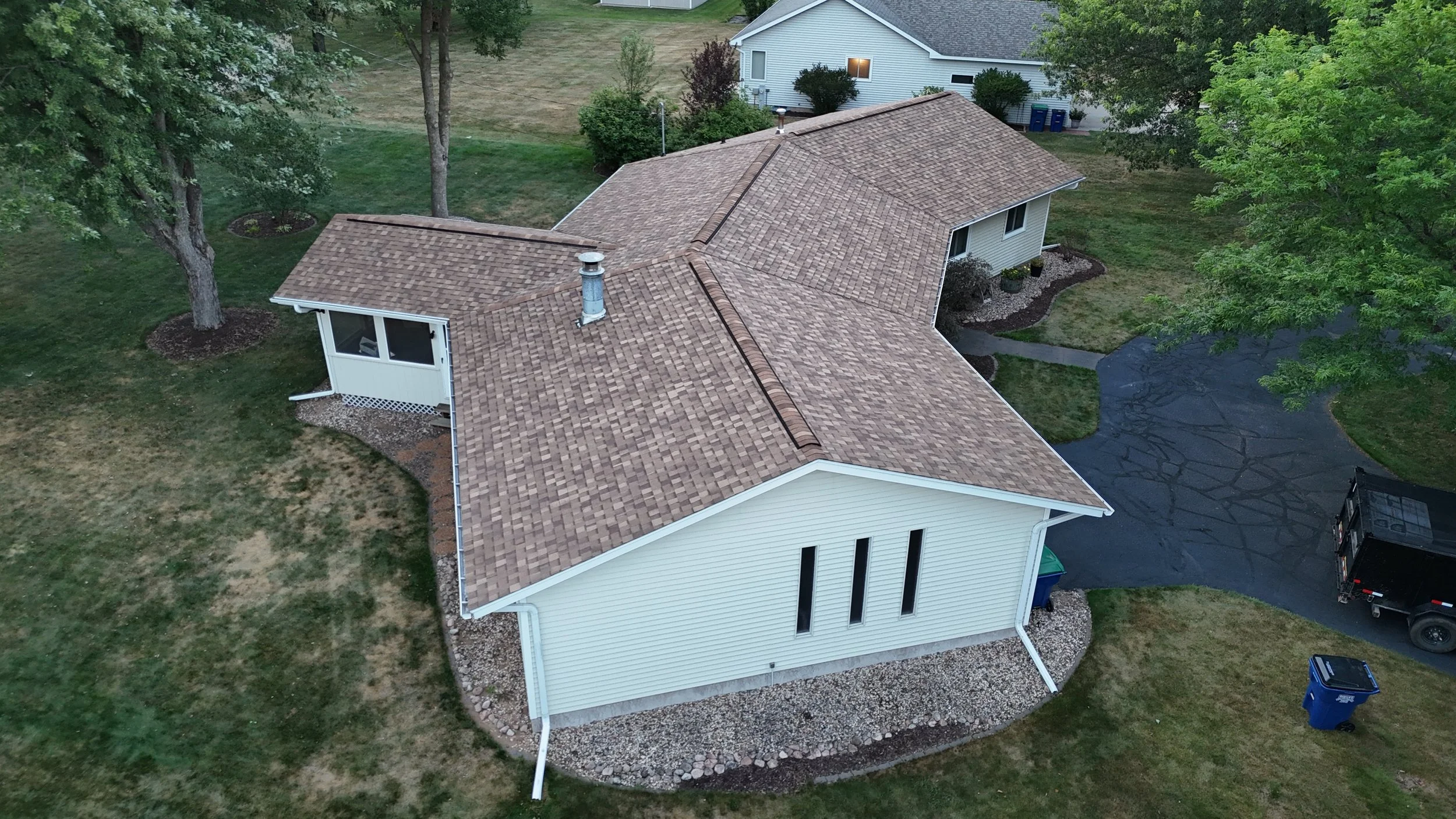 Aerial view of a house with a brown shingle roof, surrounded by green trees and grass, with a black asphalt driveway and a blue recycling bin nearby.