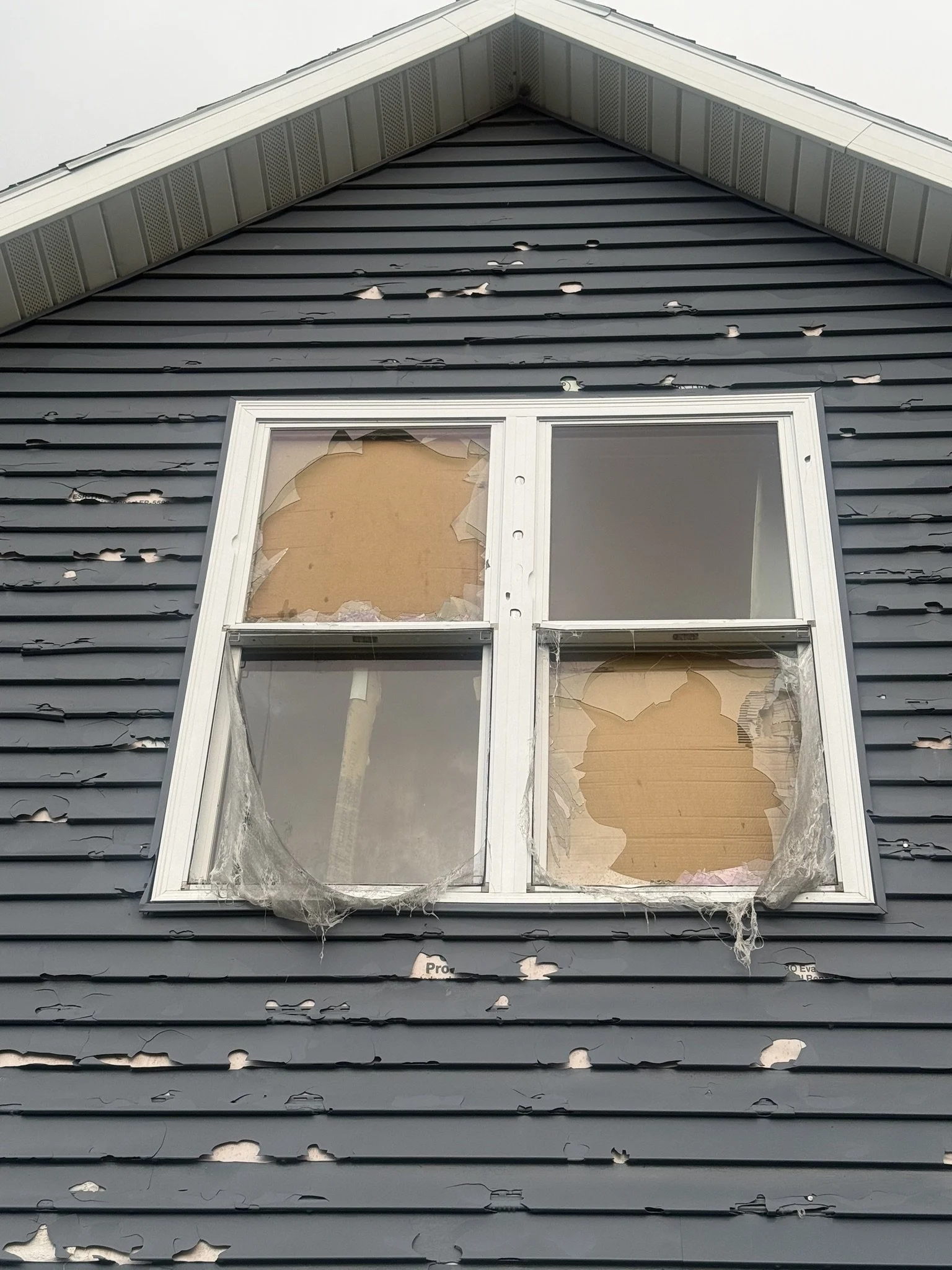 A house with damaged, black siding and a broken window in the upper level. The siding is peeling and cracked, and the window is missing glass with torn plastic around the frame.