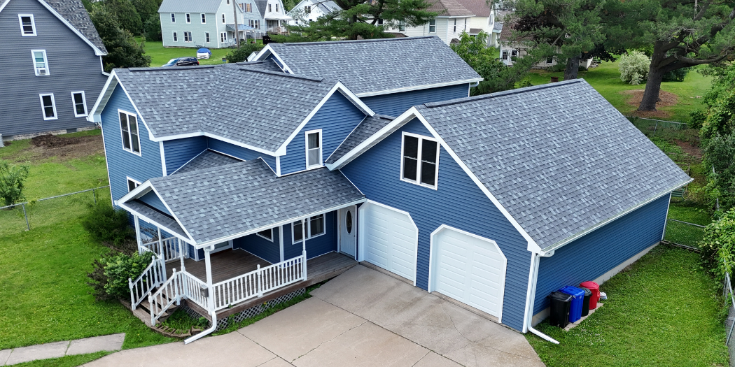 A blue two-story house with a attached garage, small front porch, and well-maintained lawn in a neighborhood with other houses and trees.