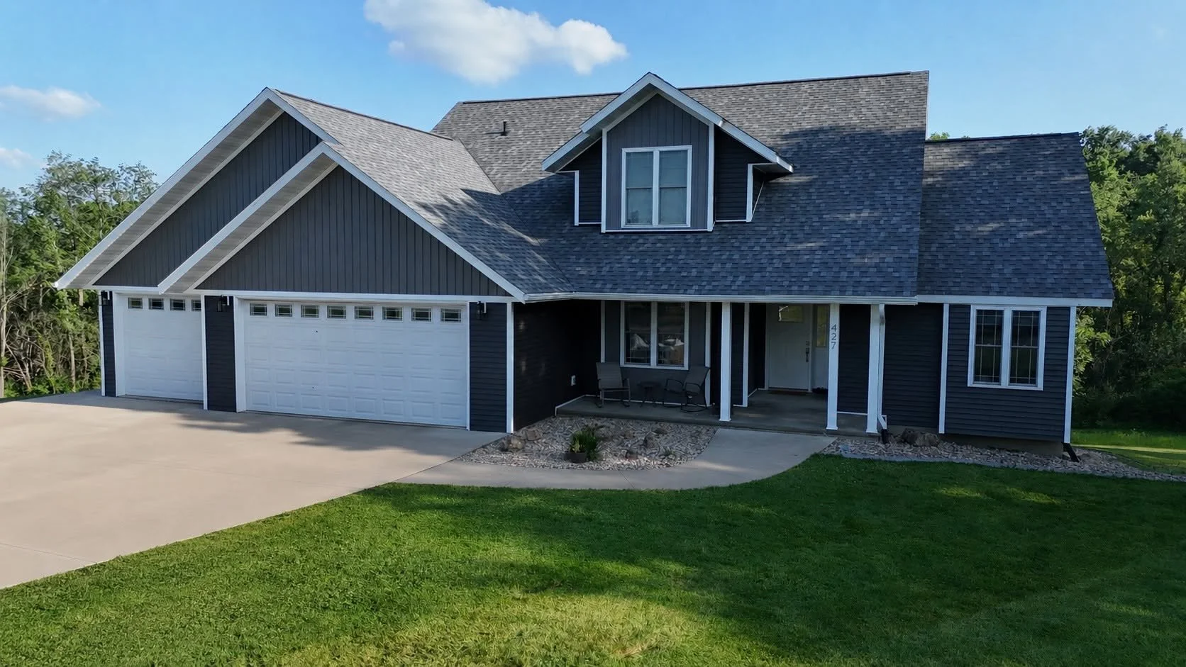A modern two-story house with a dark blue exterior, white trim, and a gray shingle roof. It features a three-car garage, a small front porch with two chairs, and a well-maintained lawn with a concrete driveway and pathway.