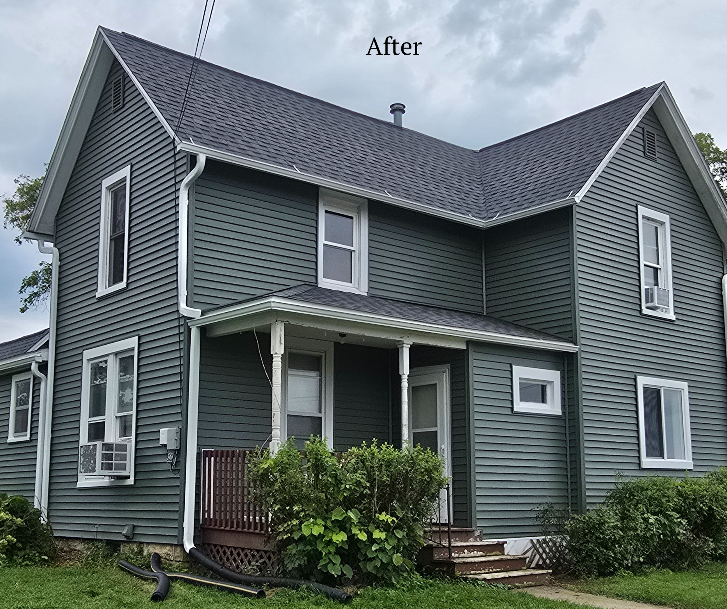 A renovated, dark green, two-story house with white trim, multiple windows, and a small front porch with steps, surrounded by green bushes and grass, under a partly cloudy sky.