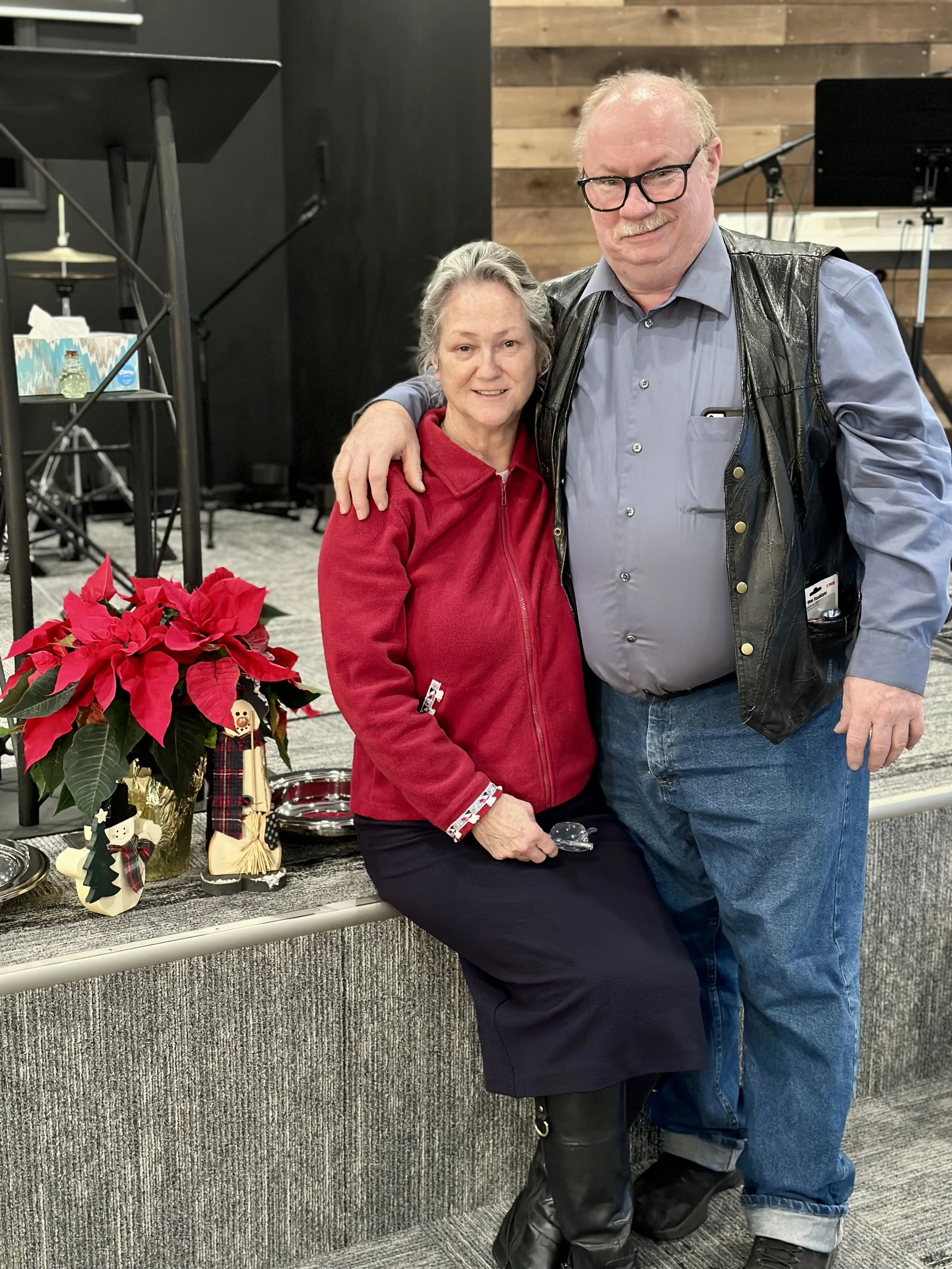 A man and a woman standing together indoors, with the man having his arm around the woman's shoulders. The woman is sitting on a ledge with Christmas decorations, poinsettia plant, and festive figurines nearby. The background has black and wooden panel walls, music stands, and a microphone stand, indicating a performance or event space.