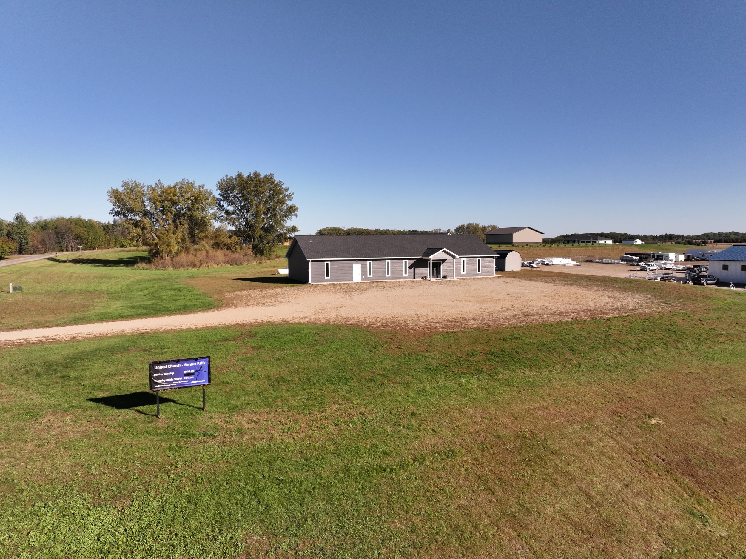 A church building with a gray exterior and black roof situated on a grassy field under a clear blue sky. There is a sign in the foreground and several other buildings and vehicles in the background.