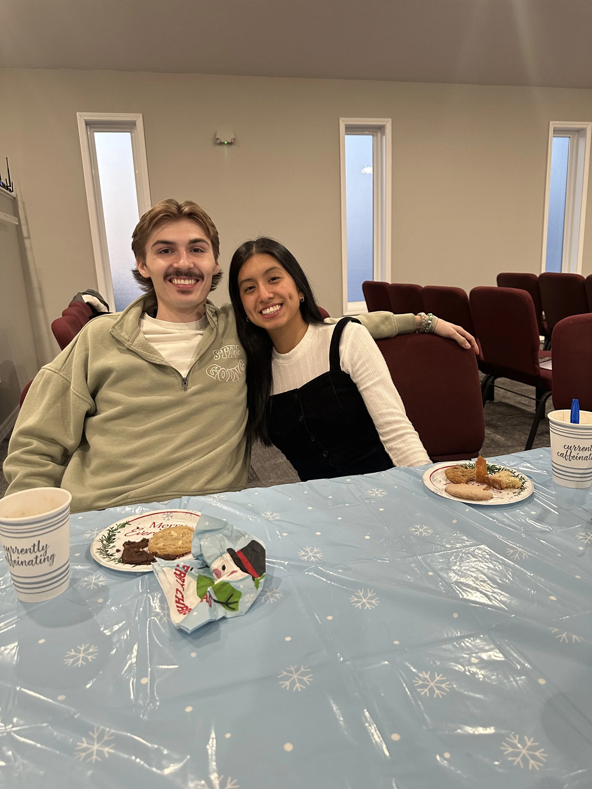 A young man and woman sitting at a table with Christmas cookies and cups, smiling and posing in a decorated indoor setting