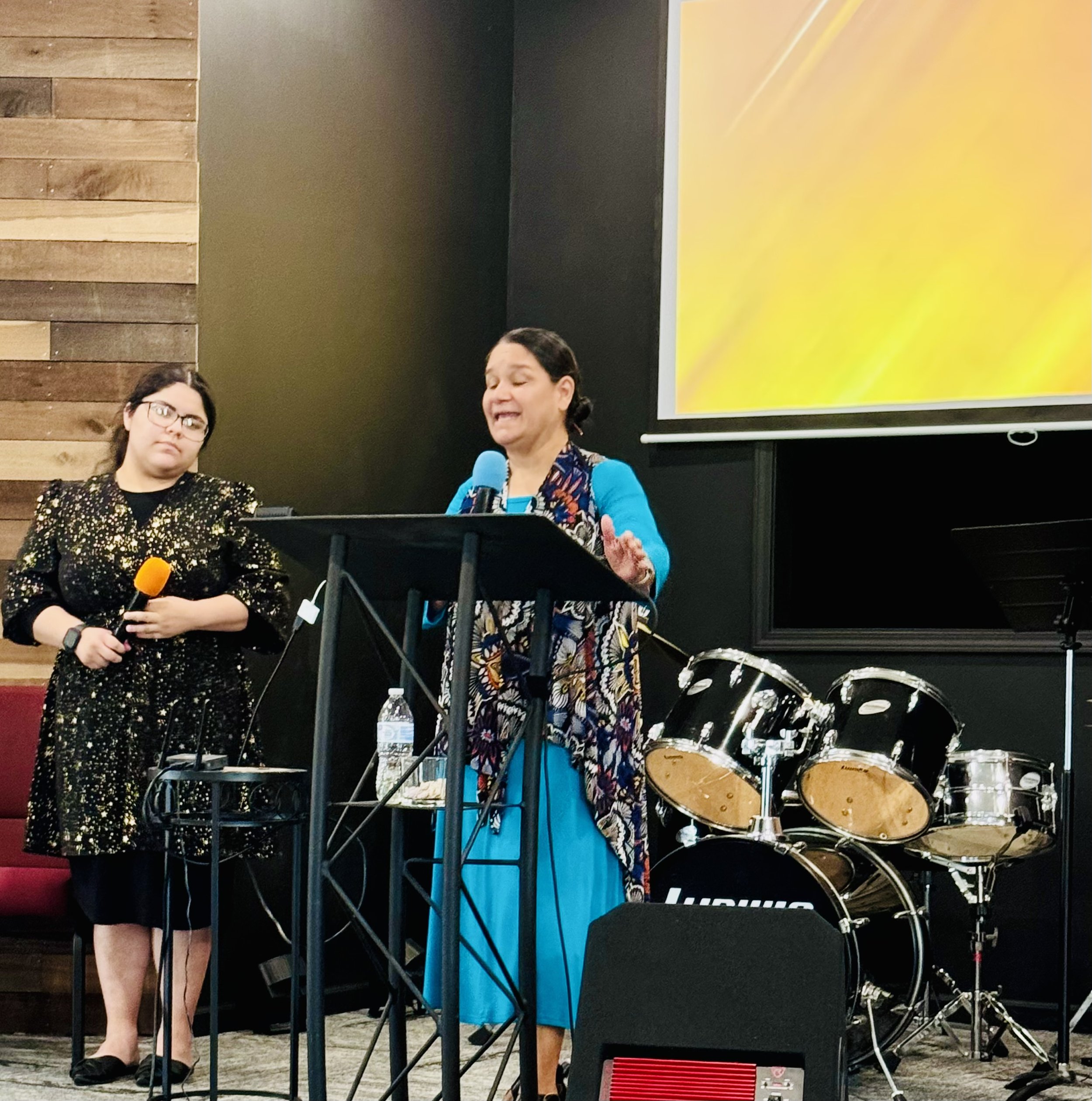 Two women speaking at a podium in a room with musical instruments. One woman is holding an orange microphone, and the other woman is speaking with her eyes closed, near a drum set and a large yellow screen.