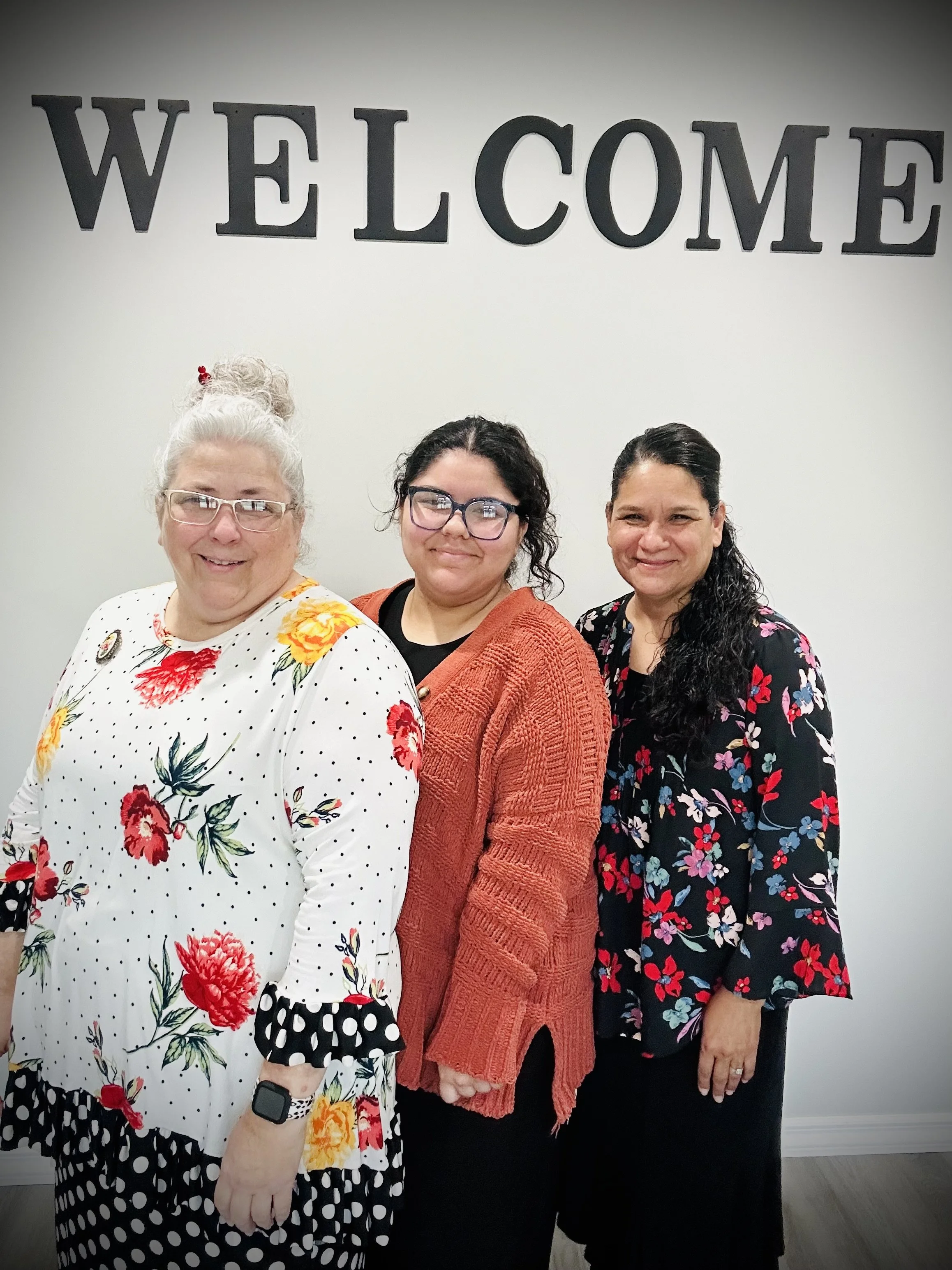 Three women standing in front of a wall with large letters spelling 'WELCOME', smiling at the camera, dressed in colorful floral clothing.