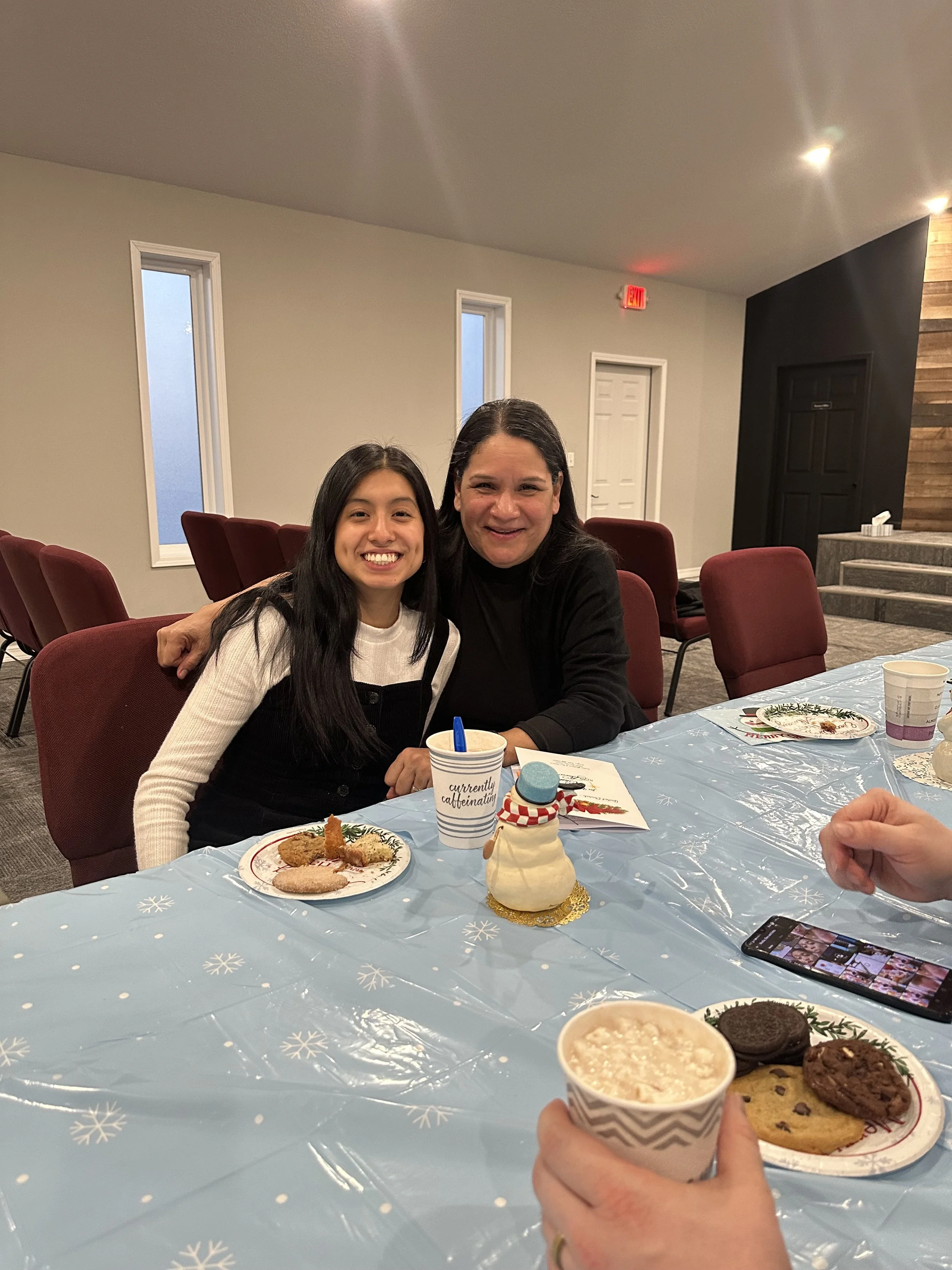 Two women sitting at a table with Christmas cookies, snowman decoration, and hot cocoa, celebrating together in a decorated room.
