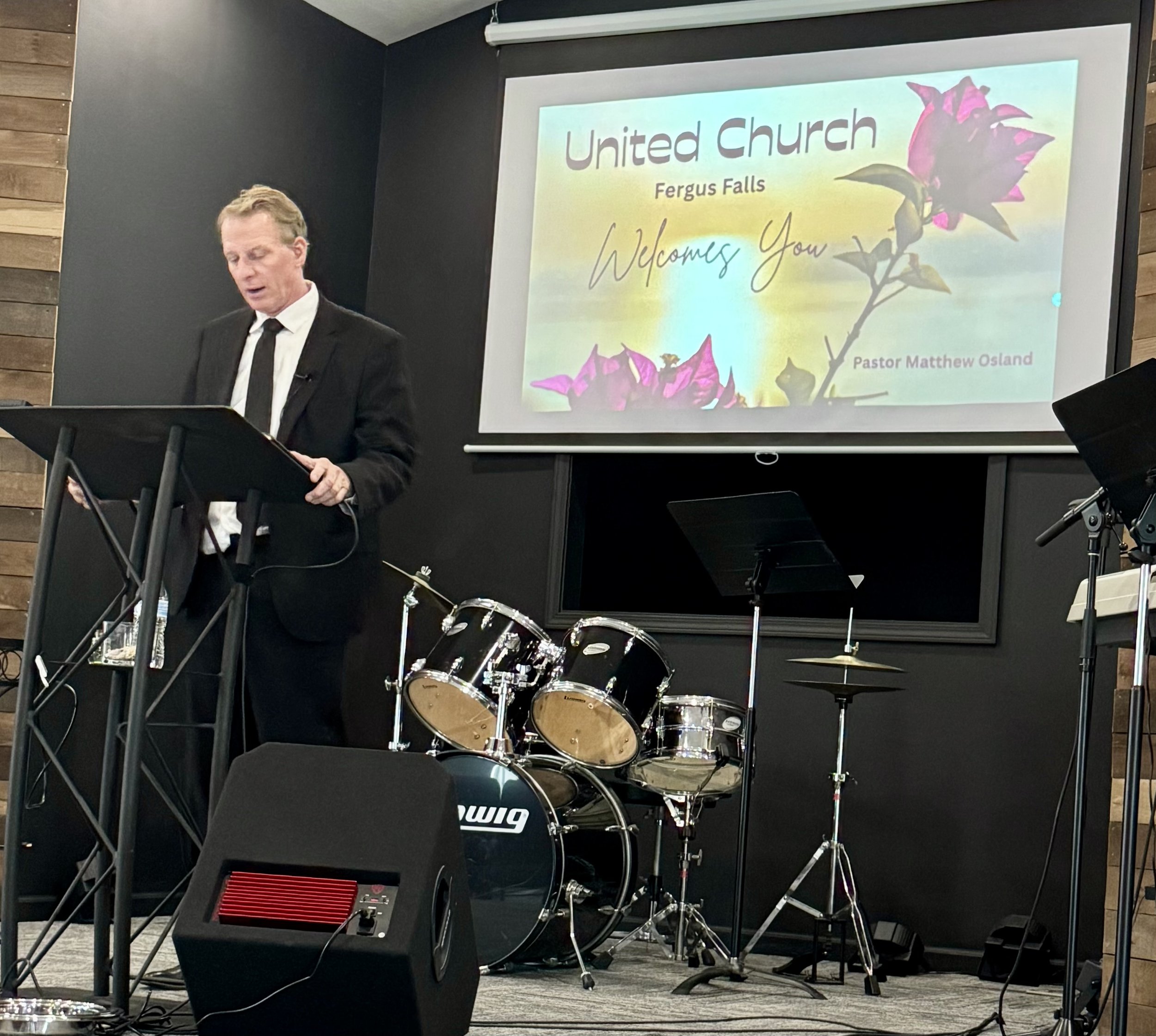 A man in a black suit and tie standing at a podium with a microphone, speaking in front of a screen that displays a welcome message for United Church in Fergus Falls, addressed by Pastor Matthew Osland.