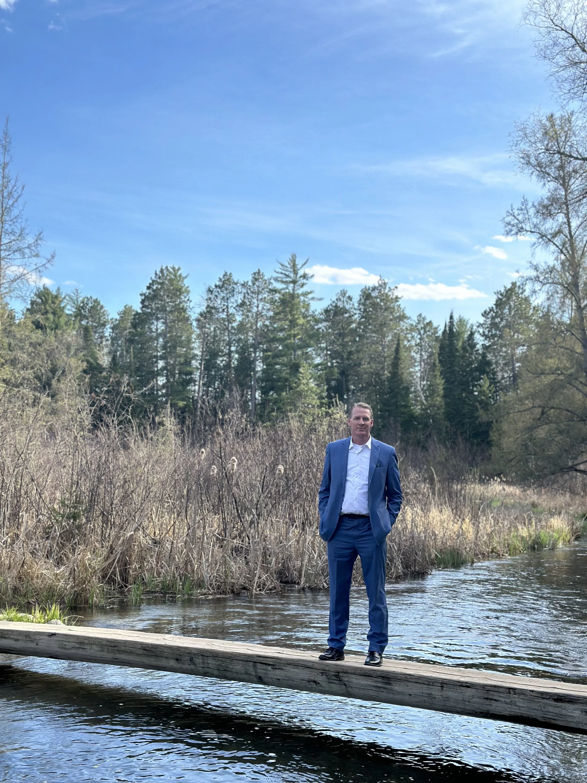 A man in a blue suit standing on a fallen tree over a river, surrounded by trees and under a blue sky.