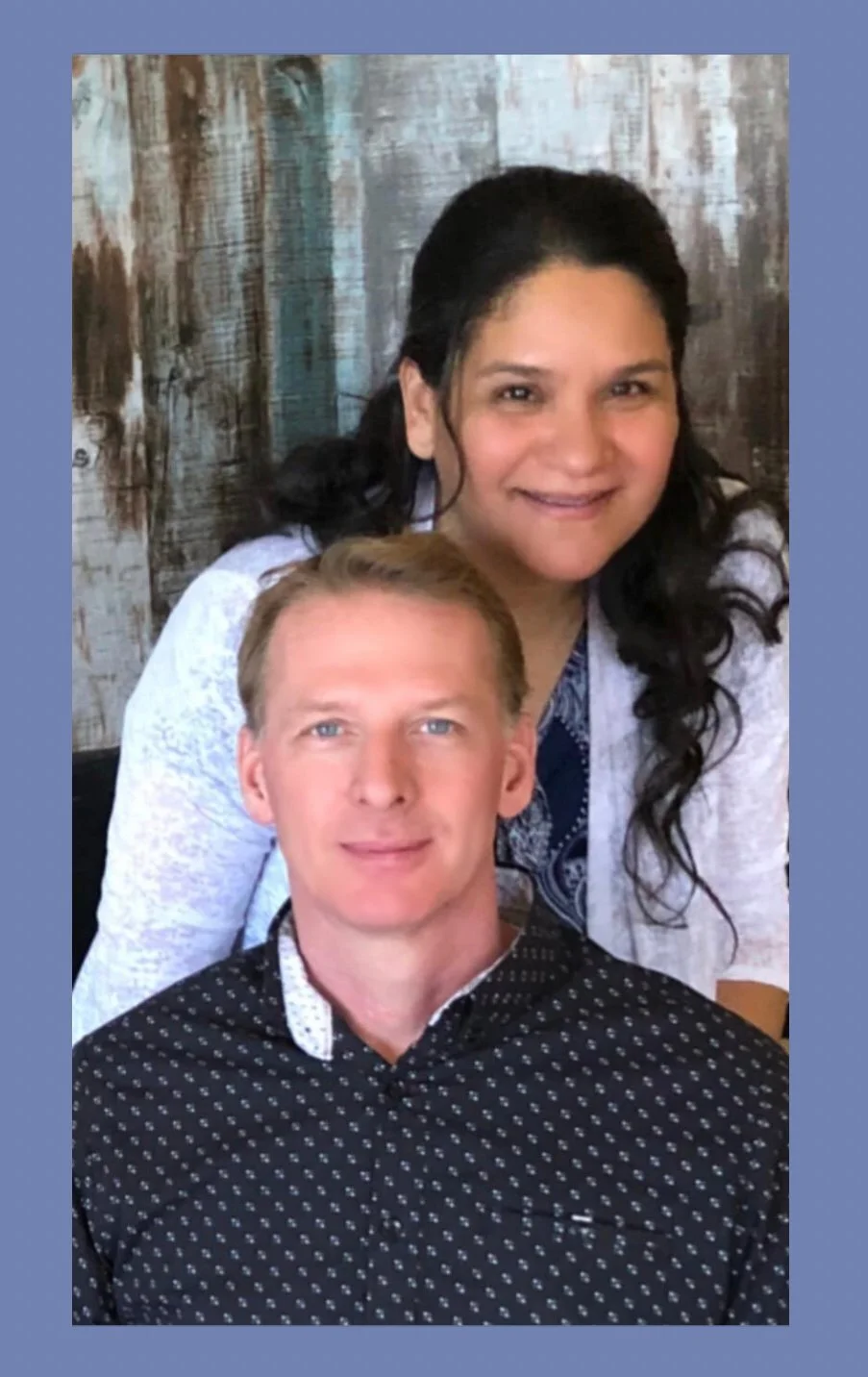 A man with blond hair and a woman with dark hair smiling for a photo. The woman is behind the man, both looking at the camera. They are indoors with a rustic wooden wall in the background.