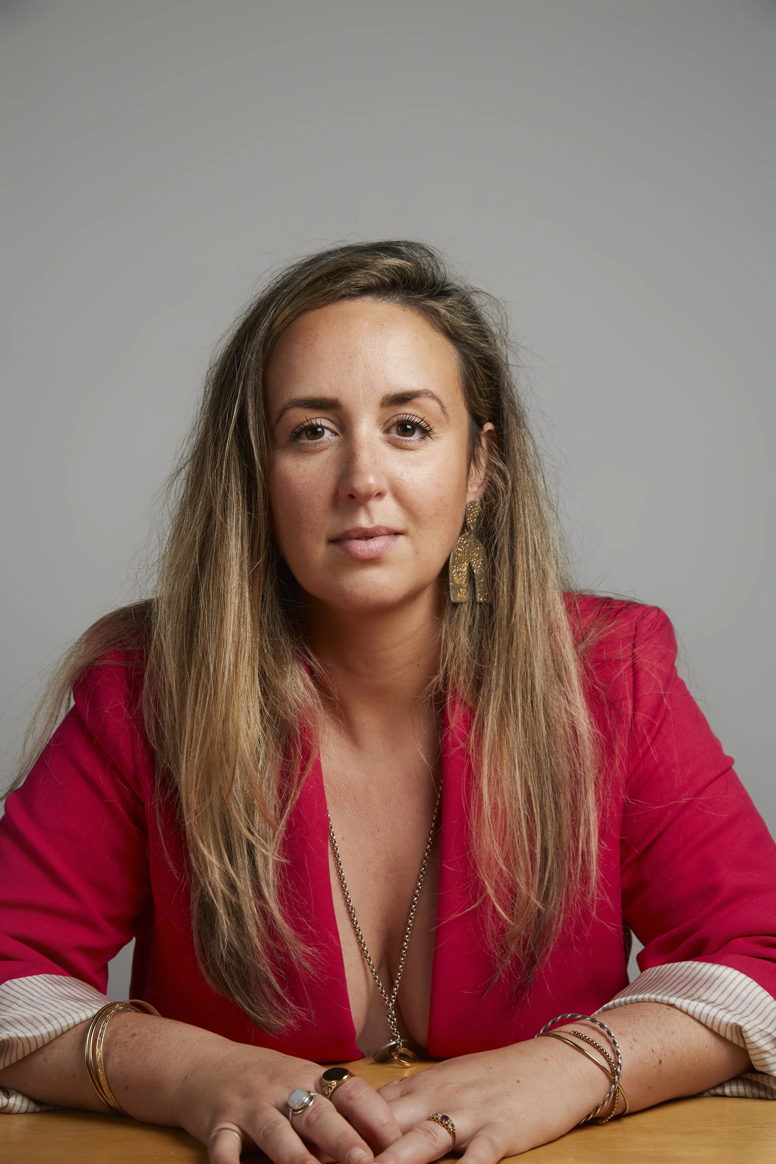 Roann Hassani McCloskey with her hair down and wearing a red blazer with a long gold chain, they are looking into the camera with kindnes, power and wisdom