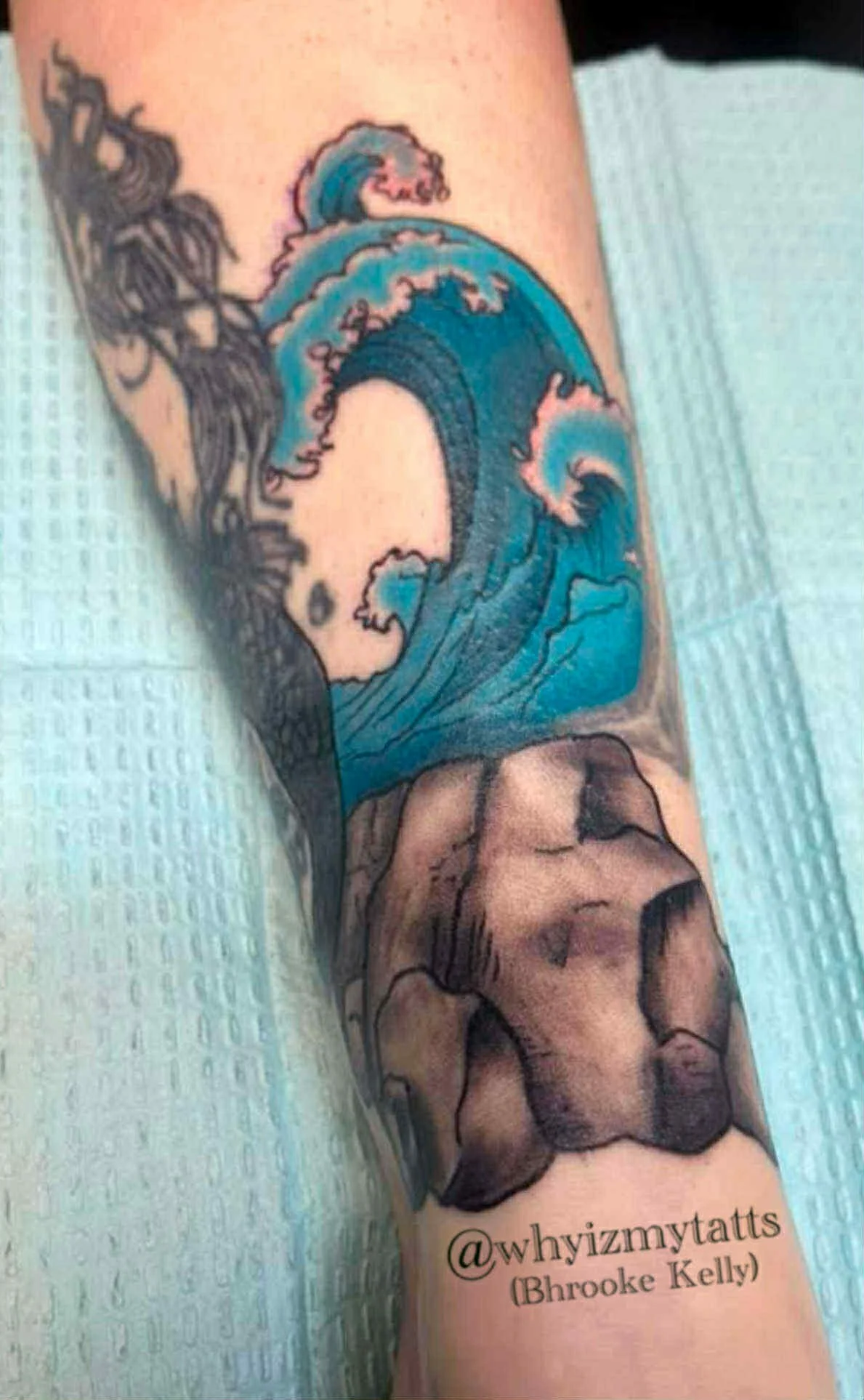 A tattoo of a large ocean wave crashing over rocks, with some black ink detail, on a person's arm. The tattoo includes a small inscription with the text '@whyizmytatts (Brooke Kelly)' at the bottom.