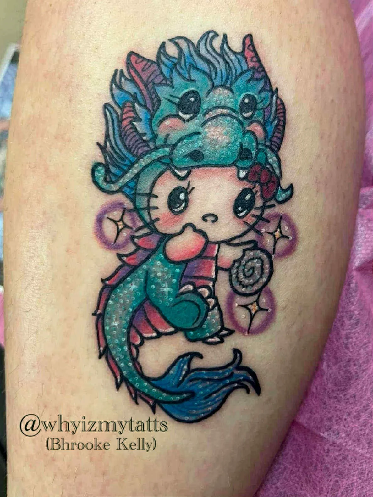 Tattoo of a cute, cartoon-style creature with a face and whiskers, combined with a dragon and a mermaid, holding a spiral shell, surrounded by purple sparkles.